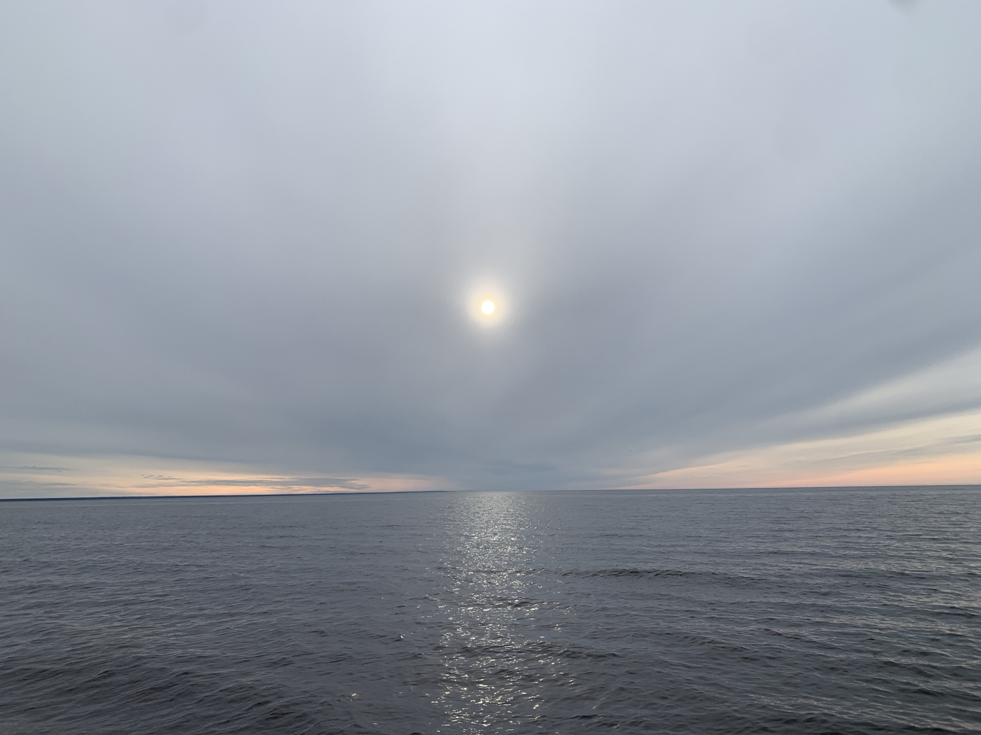 The image depicts a vast body of water, likely a sea or a large lake, under an overcast sky. The sun is visible through the cloud cover, emitting a bright, diffused light. The water surface is slightly rippled, reflecting the sunlight in a shimmering path towards the viewer. In the distance, a faint line of land is visible on the horizon. The overall color palette is muted, with shades of gray, blue, and white dominating the scene. There are no people or other objects clearly visible in the image. The scene's location is likely in the city of Severodvinsk, Russia, based on the northern latitude and coastal environment.