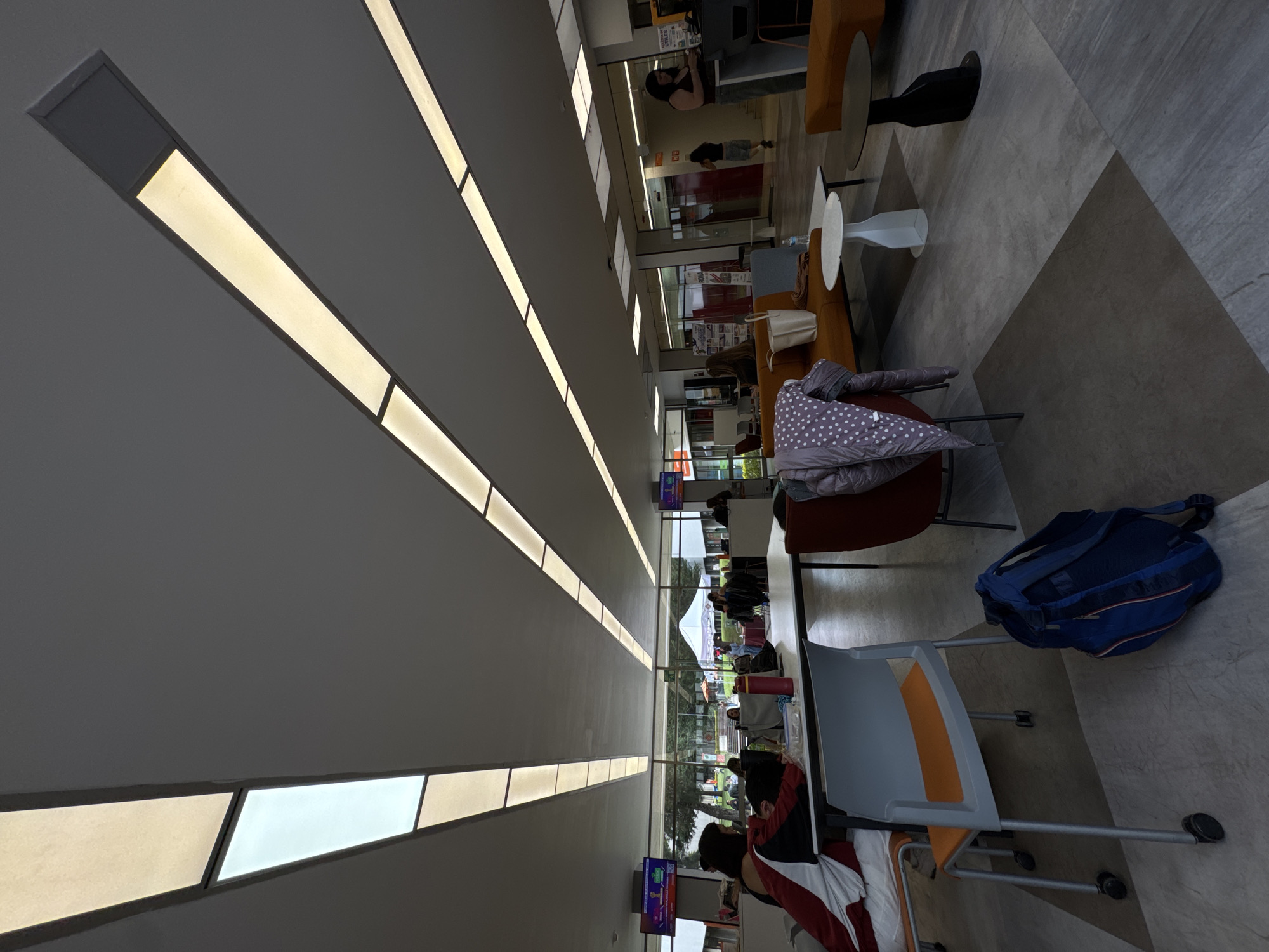 This media file depicts a modern, brightly lit indoor common area, likely a lounge or public waiting space within a building in Huixquilucan de Degollado, Mexico. The high ceiling features a distinctive design with numerous long, rectangular light panels running in parallel, providing ample illumination.
The space is furnished with various seating arrangements, including tan-colored sofas, dark red chairs, and grey chairs with orange accents, positioned around small circular side tables with white or black bases. Several people are present, mostly seated. In the foreground, a person wearing a red and white jacket is seated at a table. A blue backpack rests on the floor next to a chair, and a white polka-dot jacket is draped over another chair. Other individuals are scattered throughout the area, appearing to be engaged in activities such as studying, waiting, or socializing.
The floor is covered with large, geometric tiles in varying shades of grey, forming an intricate pattern. Large windows along one side of the room offer a view of an outdoor area, revealing green trees and a white canopy or tent, suggesting an external event or open space. Small digital screens are mounted throughout the room, displaying colorful graphics and text, with one clearly visible screen showing Spanish words like "ENFOQUE," "TALLERES," and "EXPERIENCIAS," indicating an event or program. The scene suggests a calm daytime environment, possibly a university campus lounge or a public access area.