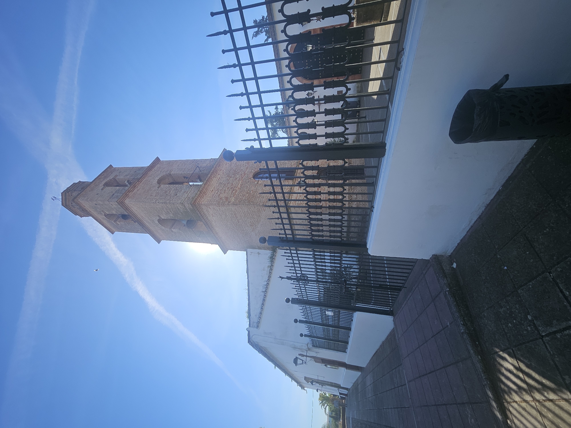 The image captures a view in Pedro Abad, Spain, featuring the brick bell tower of a church against a clear blue sky. The tower is illuminated by sunlight. A white building with a dark metal fence runs parallel to the tower. Part of a tiled walkway is visible at the bottom of the image. The sky shows contrails from aircraft.