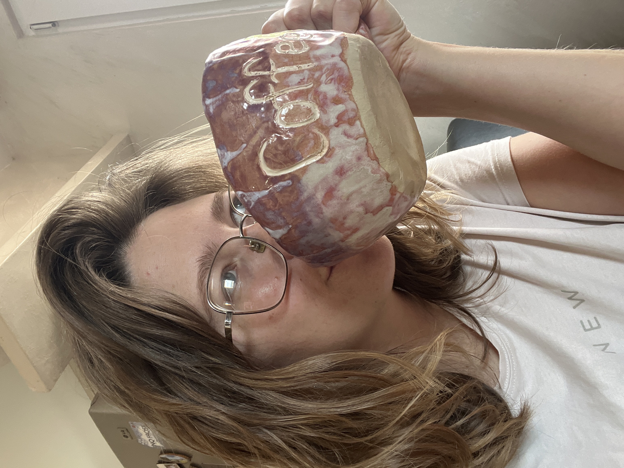 A woman is holding a large, handcrafted ceramic mug close to her face, almost touching her lips. The mug is predominantly brown and purple, with a lighter tan or cream-colored side. The word "Colu" is inscribed on the mug in a light-colored, possibly white, clay. The woman is wearing glasses with a thin metal frame. She has shoulder-length brown hair with blonde highlights. She is wearing a light pink or white t-shirt. The background includes a light-colored wall, a window, and a refrigerator with magnets on it. The location is not discernible within the image, so any reference to Podgorica, Montenegro is unsupported.