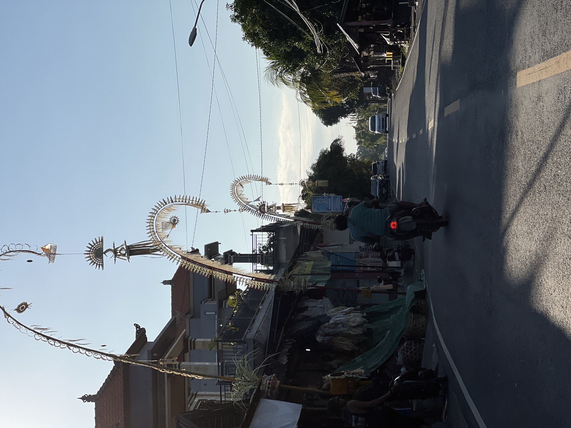 A street scene in Kubu, Indonesia, features traditional Balinese decorations arching over the road. These decorations, known as "penjor," are made of woven palm leaves and other natural materials, and are often erected during religious festivals. The scene is captured in the early morning light, casting long shadows across the asphalt. A lone motorcyclist is seen riding away from the camera, heading down the road. Several parked cars and a few buildings line the street, suggesting a small town or village setting. The sky is clear blue, with a few wispy clouds. The overall impression is one of a peaceful and vibrant community, with a strong connection to its cultural heritage.