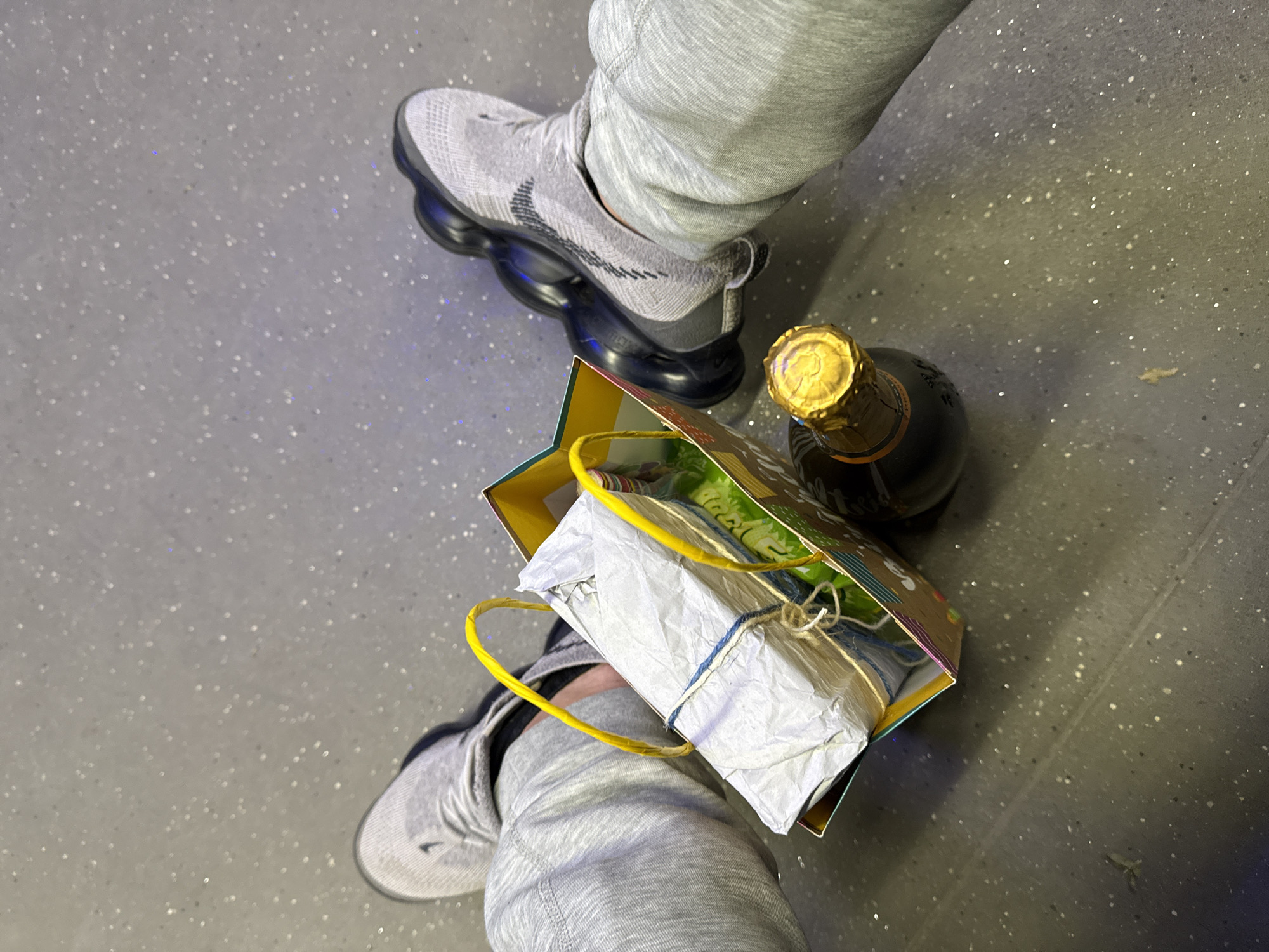 An overhead shot shows a person wearing grey sweatpants and grey Nike shoes, standing on a speckled grey floor. A gift bag with yellow handles is between the person's legs, holding a white-wrapped package tied with twine. Next to the gift bag is a dark bottle with a gold foil top, likely champagne or sparkling wine. The location is unknown and cannot be deduced to be the city of Sofia, Bulgaria from the image alone.