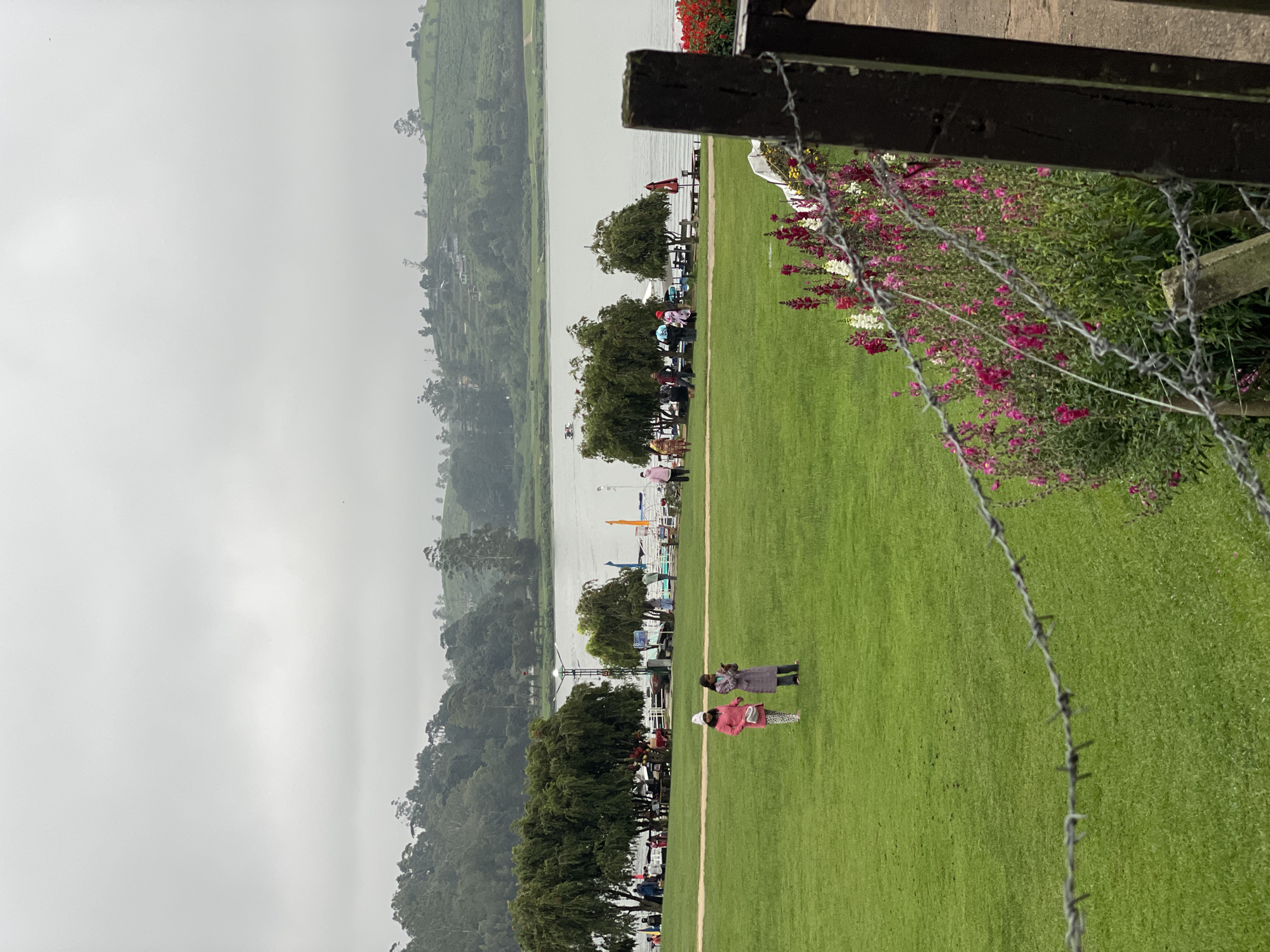 The image shows a landscape scene in Nuwara Eliya, Sri Lanka. In the foreground, there is a green lawn with two people on it. A barbed wire fence and wooden structure are partially visible on the right side of the frame. A line of trees separates the lawn from a body of water. Across the water, there are hills covered with trees and possibly tea plantations, fading into a misty, overcast sky. There are a few small structures and what appear to be docks along the water's edge. Some people are near the water. The scene conveys an impression of a cloudy, somewhat cool day.
