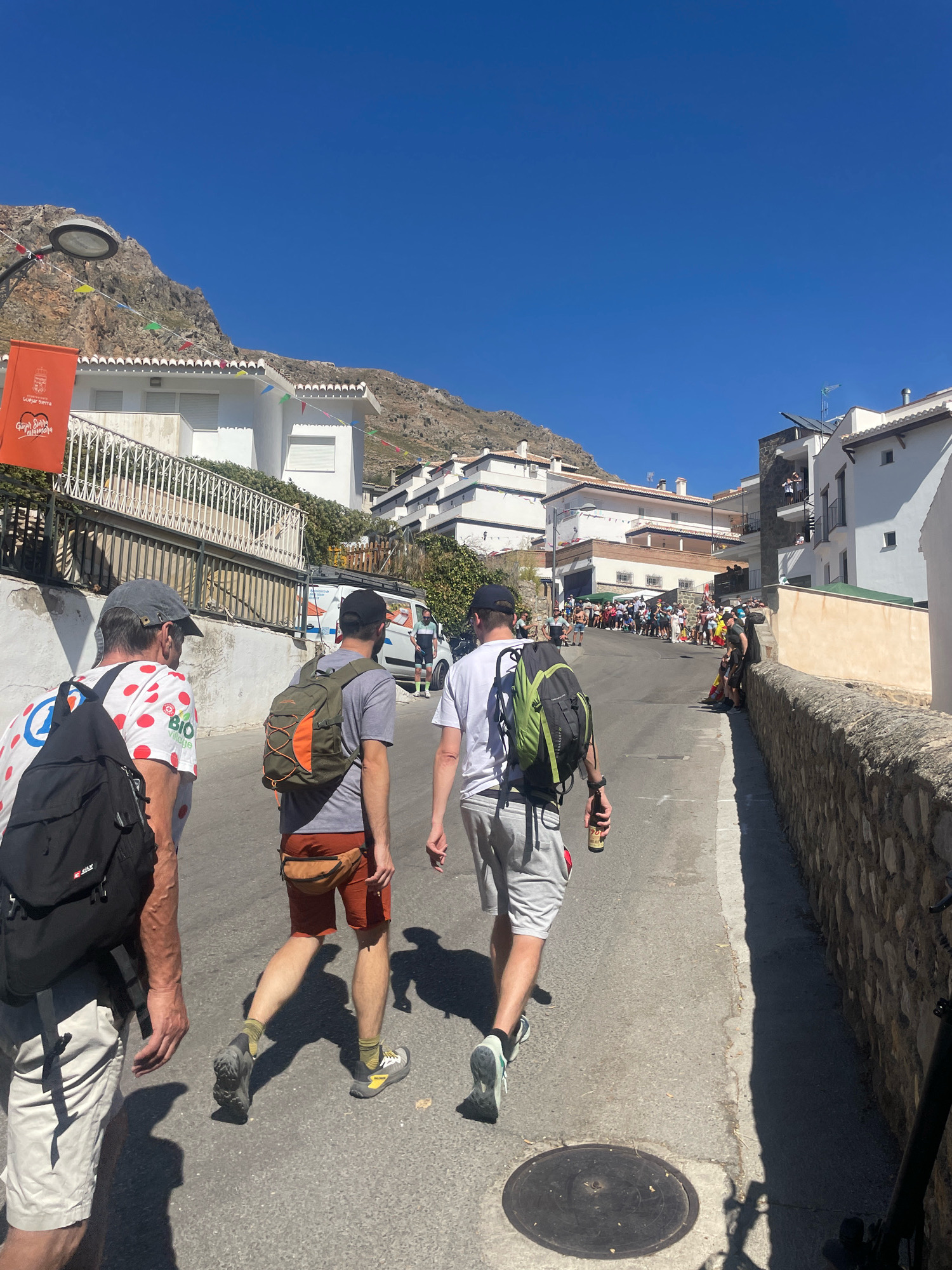 In Güéjar Sierra, Spain, three men are walking up a narrow, paved street lined with white buildings and a stone wall. The sky is clear and blue. The man in the foreground, on the left, is wearing a white t-shirt with red polka dots, a grey baseball cap, and carrying a black backpack. He is wearing light-colored shorts. To his right, a man in a grey t-shirt and orange shorts walks with an olive green backpack, a brown fanny pack, and grey athletic shoes. He is holding a can in his right hand. The third man, on the right, wears a white t-shirt and grey shorts, also carrying a backpack. In the background, a crowd of people lines the street, and there are more buildings on the hillside. A banner hangs on a building on the left, featuring a crest and text. Decorations, including small colorful flags, are strung across the street and buildings.