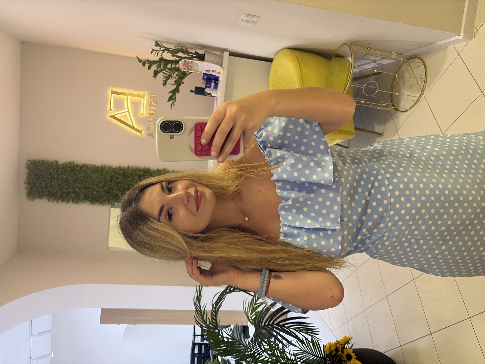A woman with long blonde hair is taking a mirror selfie in a salon or spa. She is wearing a light blue dress with white polka dots. The dress has puff sleeves and a sweetheart neckline. She is wearing a delicate necklace and bracelets on her left wrist. The background shows a reception area with a sign that reads "FA ESTETIC". There is a bright yellow chair, a gold and glass display stand, and some plants. The overall impression is of a modern and stylish establishment. The lighting suggests it is daytime.