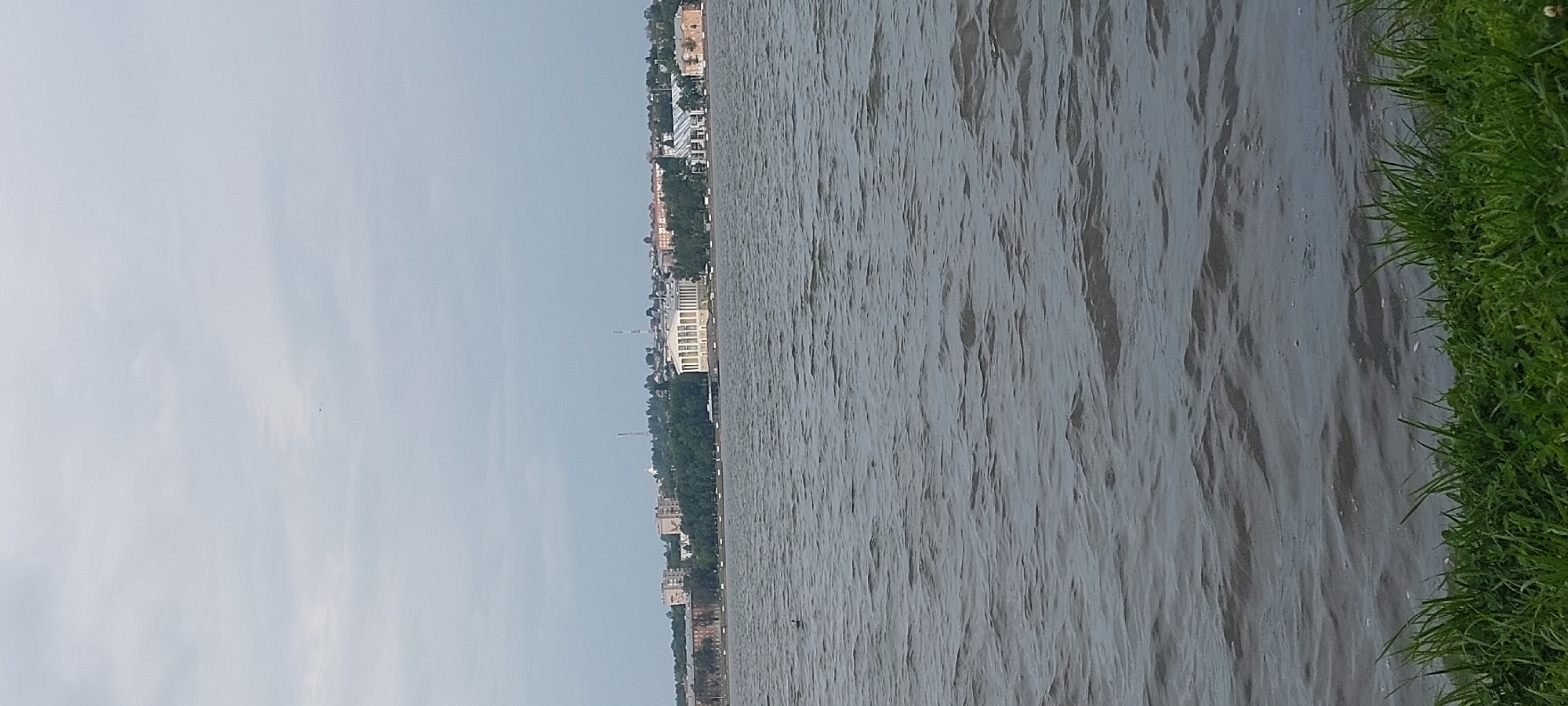 The image depicts a view of the Kama River in Votkinsk, Russia, on a cloudy day. In the foreground, lush green grass lines the riverbank. The middle ground is dominated by the rippling surface of the river, with small waves and some whitecaps suggesting a breeze. A single duck is visible on the water. In the background, across the river, is the Votkinsk embankment, featuring a grand, light-colored building with a colonnade, likely a cultural institution, situated among trees and other urban structures. The skyline is dotted with buildings of varying heights and architectural styles, with a few tall towers or antennas visible. The sky is overcast with soft, diffused light.