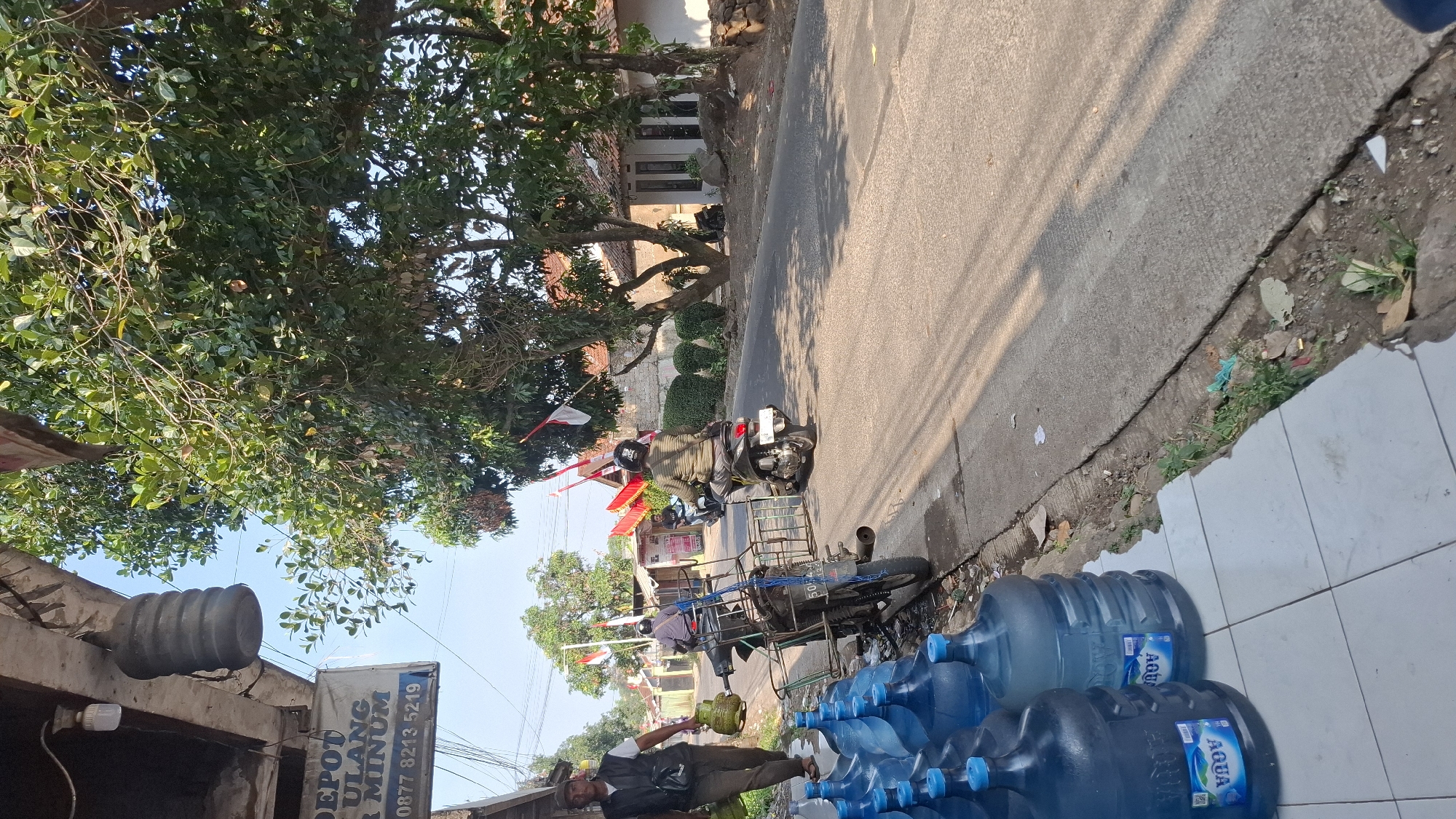 The image depicts a street scene in Cilaku, Indonesia, likely near a water depot. In the foreground, numerous large blue water bottles are stacked on a tiled surface. To the left, a man in a dark vest and brown pants walks away from the viewer, carrying a green propane tank. A motorcycle with a sidecar, also carrying a propane tank, is parked beside the water bottles. The sidecar is equipped with a metal frame. Further down the street, a person on a black motorcycle with a rider wearing a striped shirt and helmet rides away from the camera. Another motorcycle is visible in the background with a rider. Red and white Indonesian flags are displayed on poles along the street. The environment includes trees with dense green foliage and a building with a tiled roof on the right side of the road. The road is paved, and a concrete curb separates it from the sidewalk area where the water bottles are placed. The sky is clear and bright, suggesting a sunny day. The scene appears to be a typical street in a residential or commercial area of Cilaku.