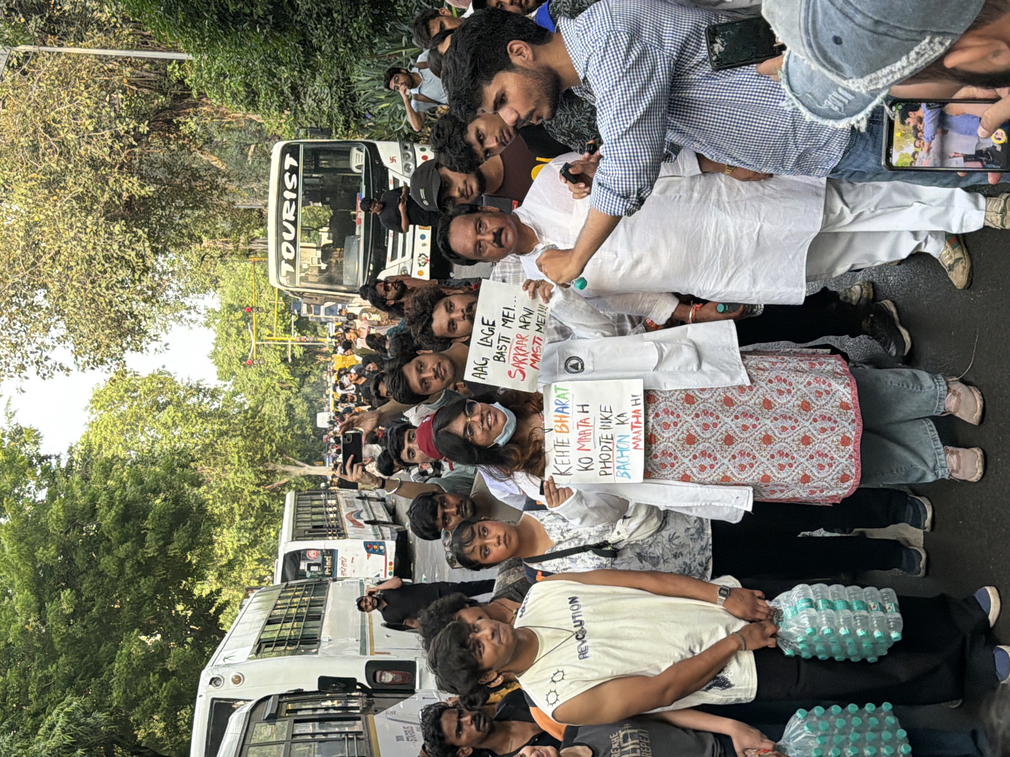 The image depicts a street scene in New Delhi, India, with a crowd of people gathered around a group holding signs. The background features several white buses, one labeled "TOURIST," and lush green trees lining the street. Traffic lights are visible in the distance, suggesting an urban environment. In the foreground, a woman wearing glasses and a white lab coat over a patterned dress holds a sign that reads "KEHTE BHARAT KO MAARTHA H PHODTE INKE BACHON KA MAATHA H!". To her right, a man in a white kurta holds another sign that says "AAG LAGE BASTI MEI... SARKAR APNI MASTI MEI!!!". Several individuals are holding mobile phones, appearing to take pictures or videos of the protest. Two people are seen carrying packs of bottled water. The overall atmosphere suggests a protest or demonstration.