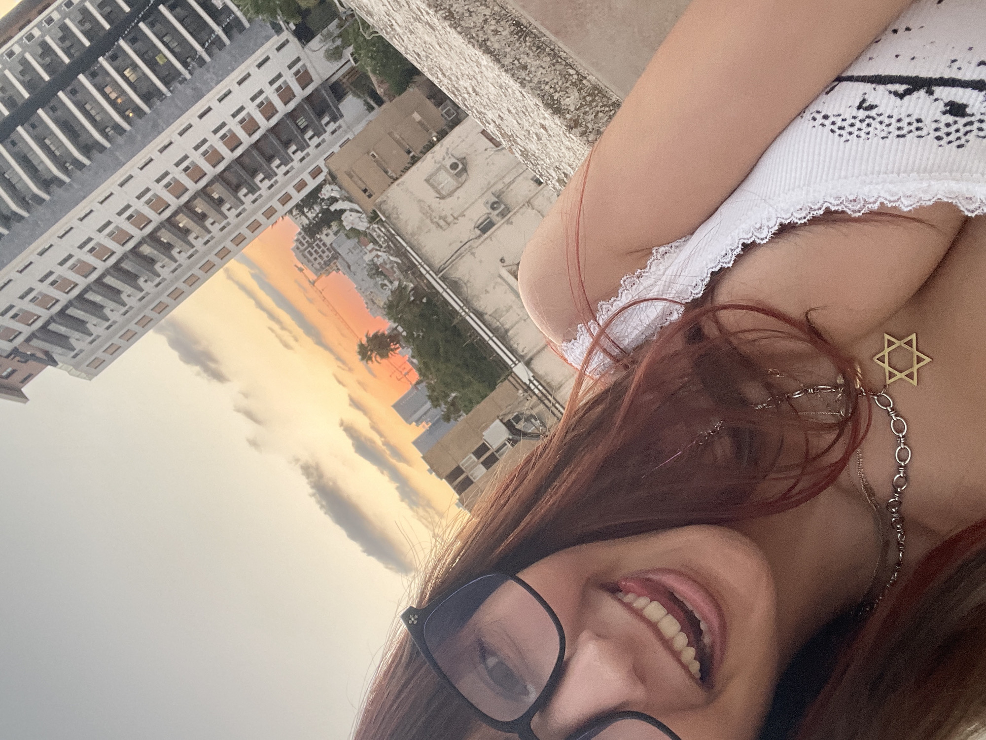 The image is a selfie taken outdoors during golden hour, with the sky displaying soft orange and yellow hues. The subject is a young woman with reddish-brown hair, wearing glasses and a white lace-trimmed top. She is sticking her tongue out and smiling. She is wearing a silver necklace with a Star of David pendant. In the background, there is a city skyline with several buildings, including a tall, modern high-rise with a gray facade and balconies. The buildings appear to be in a dense urban environment. The perspective suggests the photo was taken from a higher vantage point, possibly a rooftop, overlooking the city. The location is Petah Tikva, Israel.