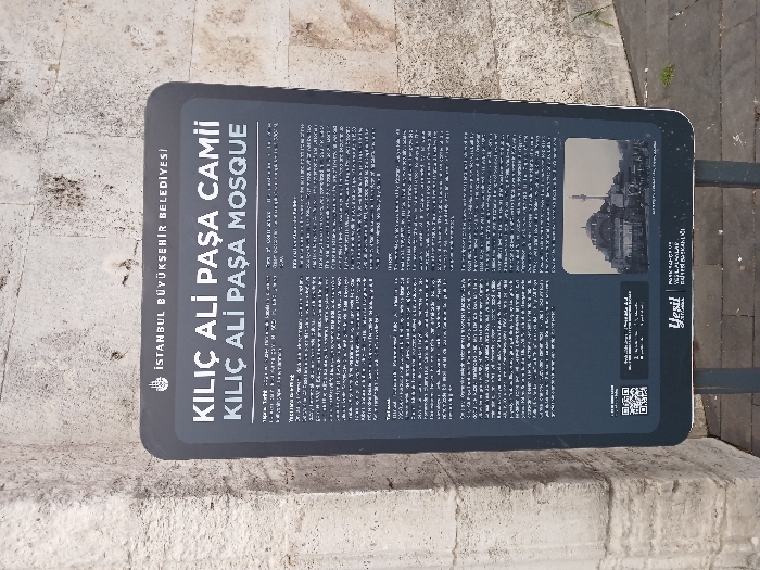 The image shows a black sign with white text attached to a white stone wall. The sign is located in Istanbul, Türkiye (latitude: 41.027, longitude: 28.98). The sign's text reads: "ISTANBUL BUYUKSEHIR BELEDIYESI KILIC ALI PASA CAMII KILIC ALI PASA MOSQUE." The sign features a QR code, two paragraphs of text, and a small black and white photograph at the bottom. The photograph depicts the mosque with a minaret, with people walking in front of it. There is a green bush in the bottom right corner. The sign is standing upright on a metal pole on a paved sidewalk.