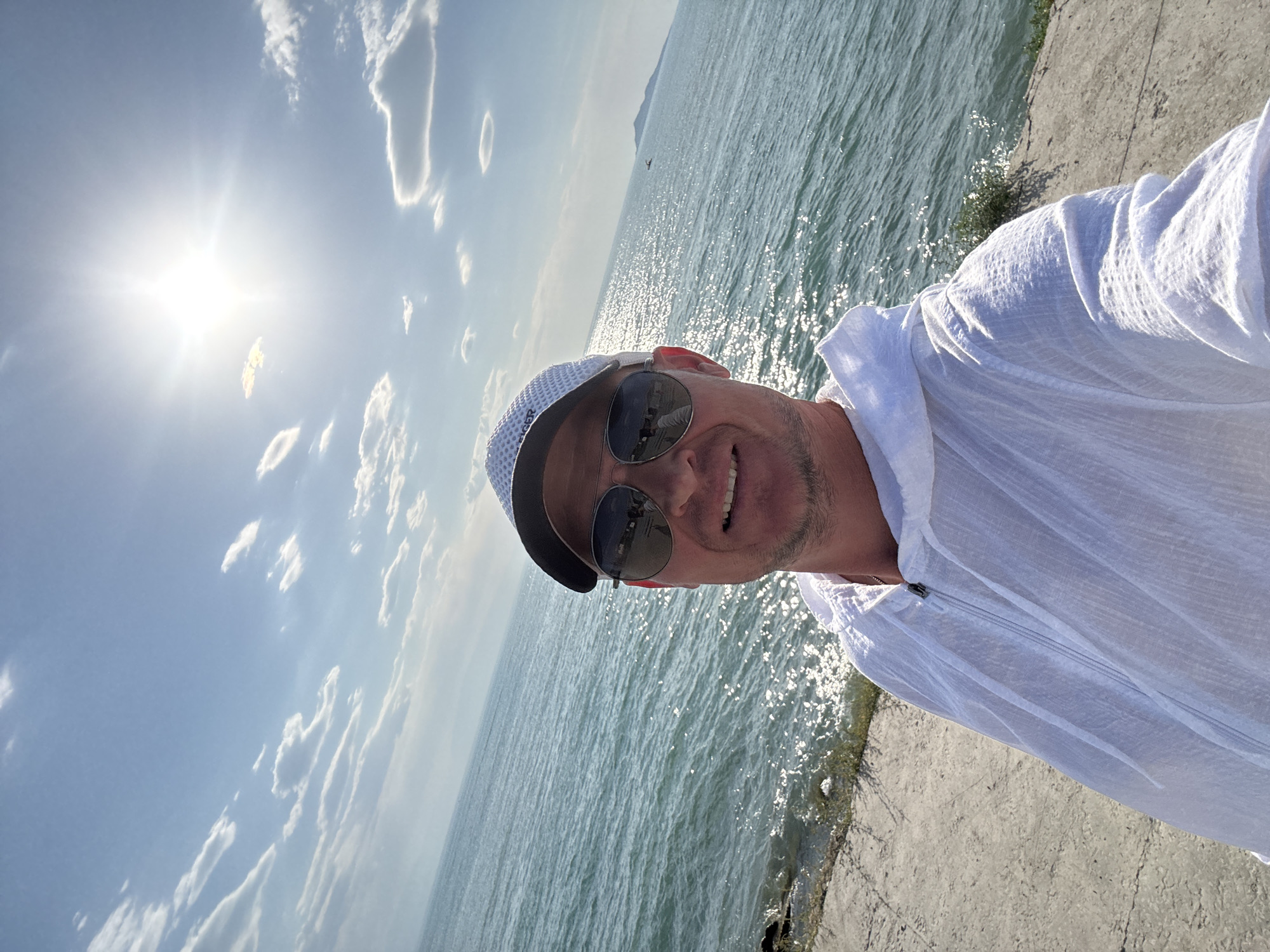 A man is taking a selfie outdoors, standing near a body of water. He is wearing a white hooded jacket, a baseball cap, and sunglasses. The sun is shining brightly in the sky, casting reflections on the water. The background shows a calm sea with some distant landmasses. The man appears to be on a paved walkway next to the water's edge. The overall scene suggests a sunny day by the water.