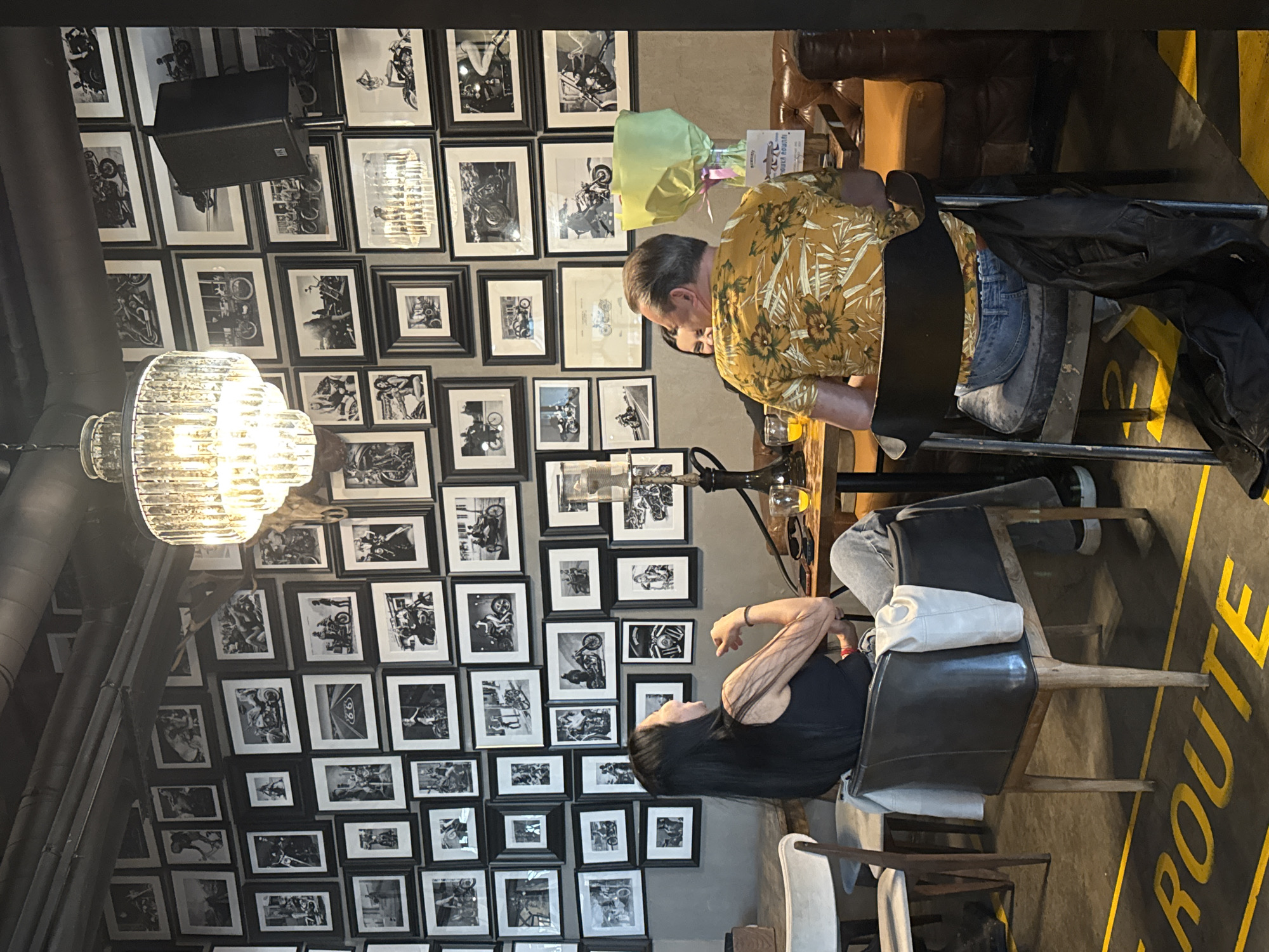 A horizontal medium-angle shot shows two people at a table in a cafe in Saint Petersburg, Russia, with a wall covered in framed motorcycle photos.
A man in a yellow floral shirt is sitting at the table facing the woman, leaning slightly forward. He has short, gray hair and appears to be talking. The woman is sitting in a black leather chair, facing away from the camera, with long, black hair and a black tank top. Her arms are crossed. On the floor next to her chair is a white bag.
Behind the table is a wall entirely covered in framed black-and-white photos of motorcycles. To the left, a large, ornate chandelier hangs from the ceiling, and a speaker system is mounted on the wall among the photos. The floor is dark with yellow painted words saying "ROUTE" on it. There are other chairs and tables visible in the background.