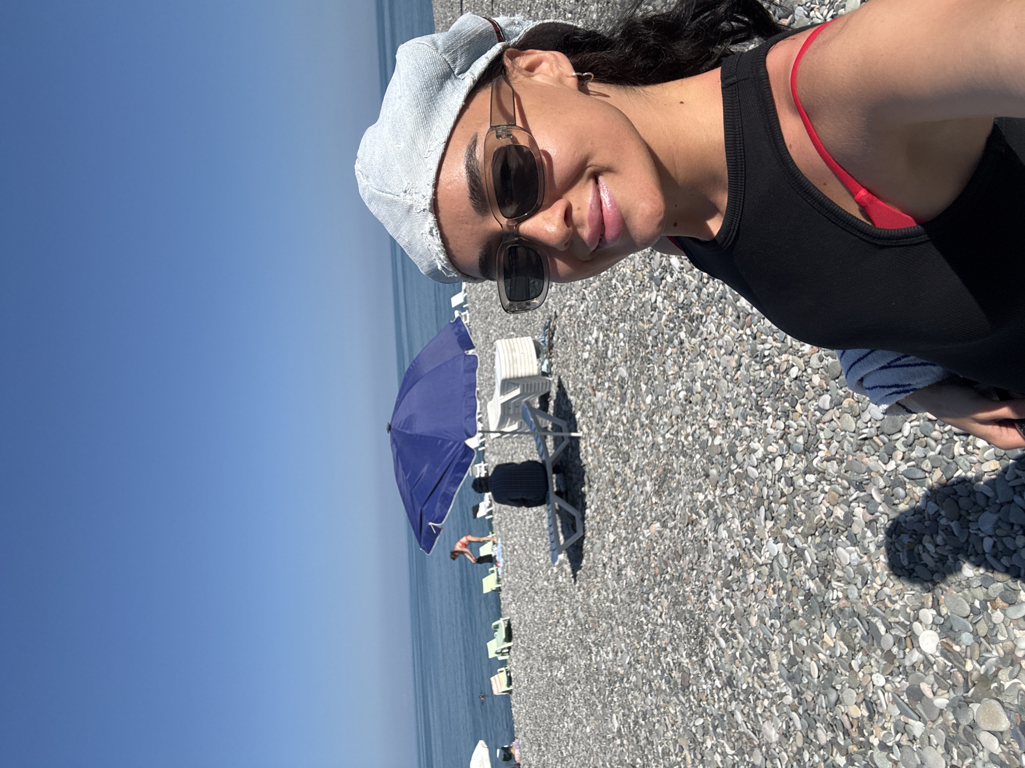 A young woman with dark curly hair and a denim cap is taking a selfie on a pebble beach in Batumi, Georgia. She is wearing sunglasses, a black tank top, and a red bikini top. The sea is visible in the background, along with several beach umbrellas and lounge chairs. A man is sitting on a lounge chair under a blue umbrella. The sky is clear and blue.