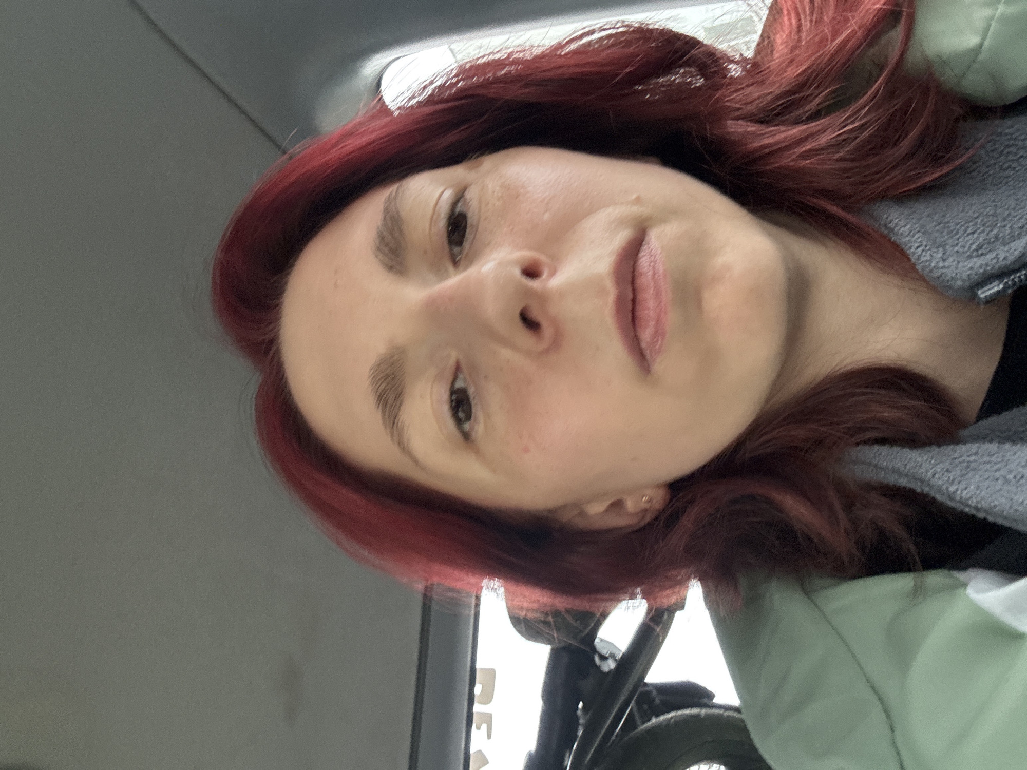A medium shot of a woman with red hair, facing the camera directly. She is wearing a green jacket and a grey scarf. The background appears to be the interior of a vehicle, with the ceiling visible at the top of the frame and a window with lettering on the left side. A bicycle is partially visible in the background on the left.