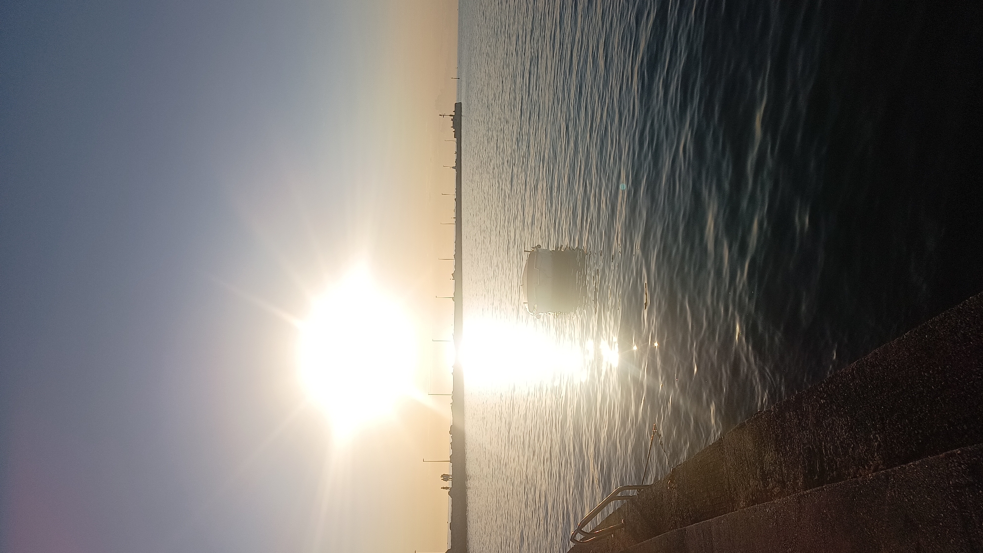 The image captures a coastal scene in Umag, Croatia, during sunrise or sunset. A prominent, low-angle sunburst dominates the upper center, generating intense glare and casting a strong, vertical reflection path on the calm, rippled water surface. In the left foreground, a weathered concrete embankment with visible steps and partial metal handrails descends into the water. A small, light-colored boat, likely white, floats in the mid-ground, positioned slightly right of the sun's reflection. A long, dark pier or breakwater extends horizontally across the mid-distance. Three to four indistinct human figures are visible on the left side of the pier, silhouetted against the bright light. Multiple evenly spaced lampposts or tall structures are distributed along the pier. At the far right end of the pier, a structure resembling a lighthouse or beacon is present. The sky above is clear, transitioning from bright white-yellow near the sun to a pale blue-grey. A faint horizon line and distant landmass are visible in the far background.