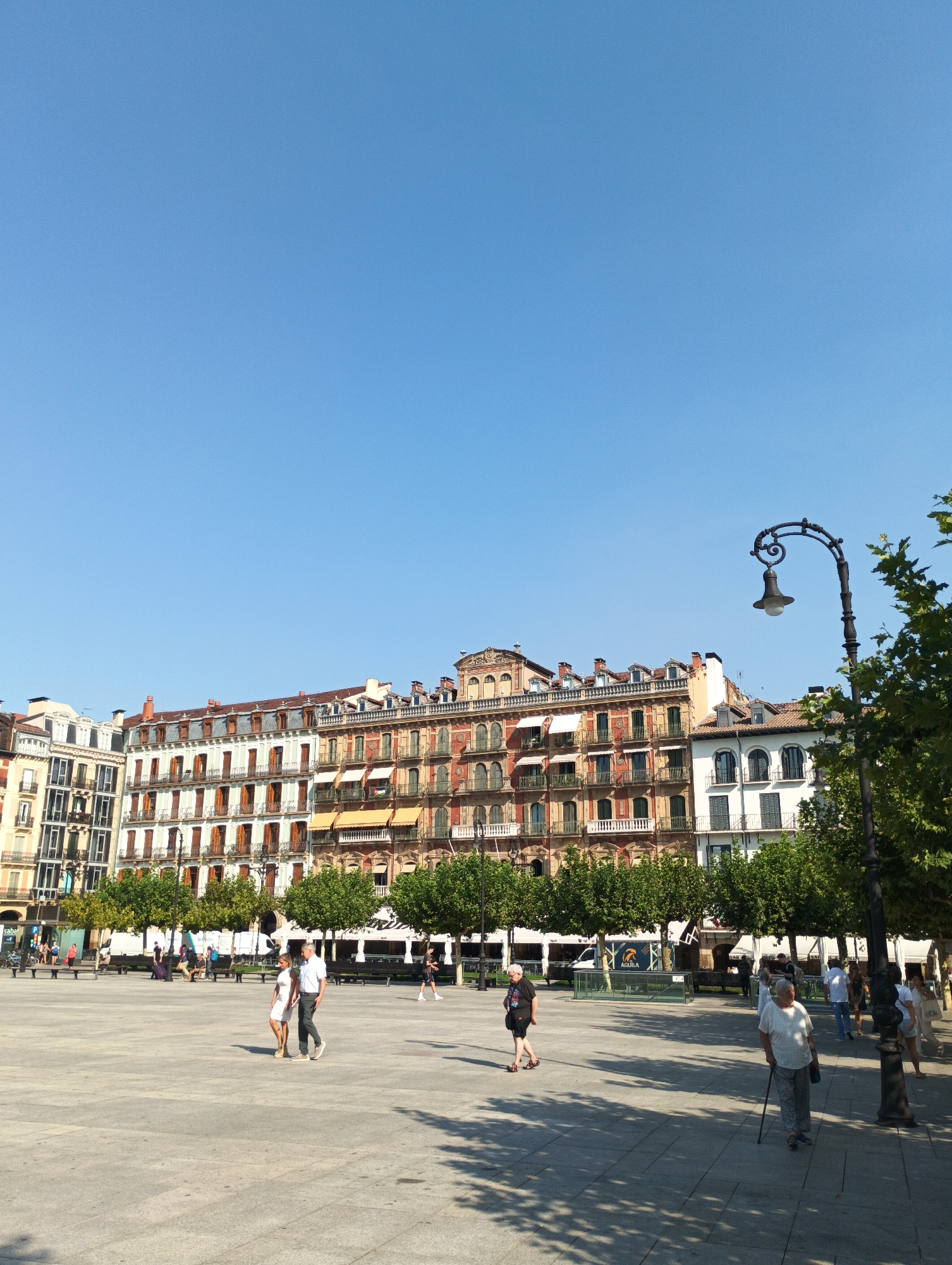The image depicts a sunlit plaza in Pamplona, Spain, likely Plaza del Castillo, given the distinctive architecture of the buildings lining the square. The buildings exhibit a mix of architectural styles, with several stories, numerous windows with shutters, and ornate balconies. The sky is a clear, bright blue, suggesting a pleasant day.
In the foreground, the paved plaza is sparsely populated. A couple, a man and a woman, are walking hand-in-hand towards the left. The man is wearing a white short-sleeved shirt and dark trousers, while the woman is in a white dress. Further to the right, an elderly woman with gray hair is walking with a cane, dressed in a white patterned top and gray trousers. Another elderly woman with short gray hair is walking towards the left, wearing a black t-shirt and black shorts. A tall, black, wrought-iron lamppost stands prominently in the mid-right of the frame, casting shadows on the ground.
In the background, more people are visible, some sitting on benches, others walking. Trees with dense green foliage are planted along the edge of the plaza, providing shade. Several white vans or trucks are parked behind the trees. The overall scene conveys a sense of public life and leisure in an urban setting.