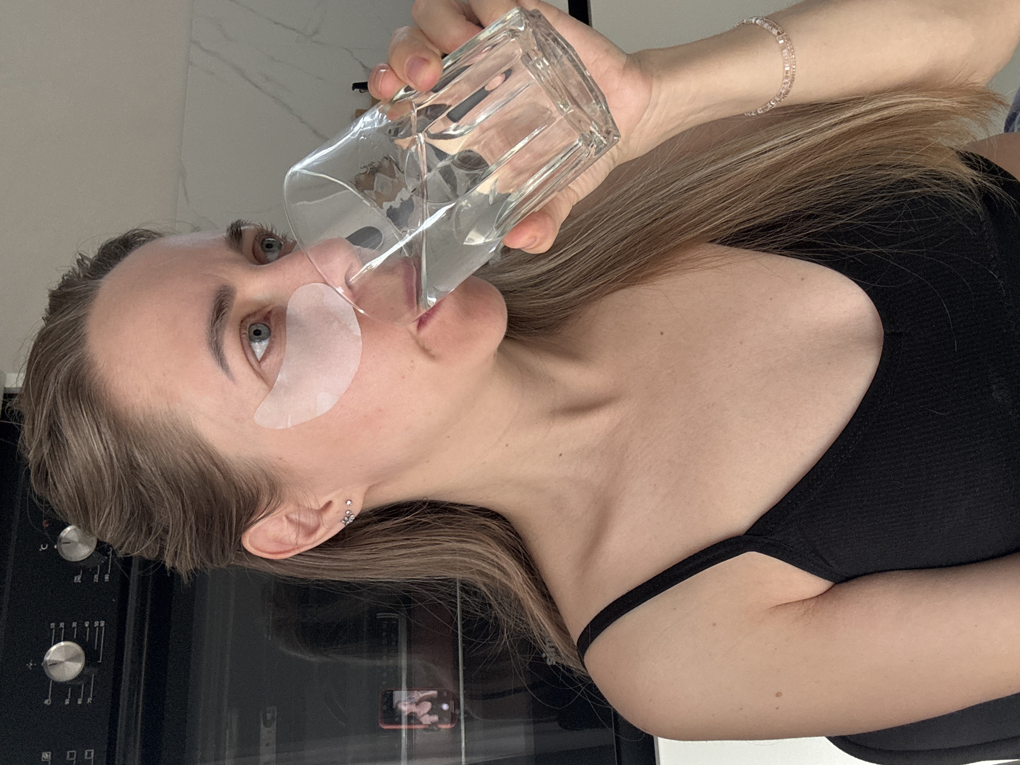A young woman, presumably in Chaikovsky, Russia, is depicted in profile, drinking water from a clear, faceted glass. She has long, light brown hair, which falls over her shoulder, and light-colored eyes. She is wearing white, gel-like under-eye patches, indicating a skincare routine. Her attire includes a simple black top, and she accessorizes with a delicate, light-colored bracelet on her right wrist and multiple small stud earrings in her left ear.
The activity taking place is the woman hydrating herself. Her expression is neutral and calm. The setting appears to be indoors, with a light-colored, possibly tiled wall in the background and dark, glossy surfaces that reflect parts of the room. A notable detail is the reflection of a mobile phone with a bright screen on one of the dark surfaces, although the content on the screen is indistinct. Below this, another dark panel features two silver knobs with abstract markings that resemble musical notes, suggesting the presence of a kitchen appliance like an oven or cooktop. The image is oriented vertically, with the woman's head tilted towards the left, capturing a moment of self-care. No legible text is visible in the image.