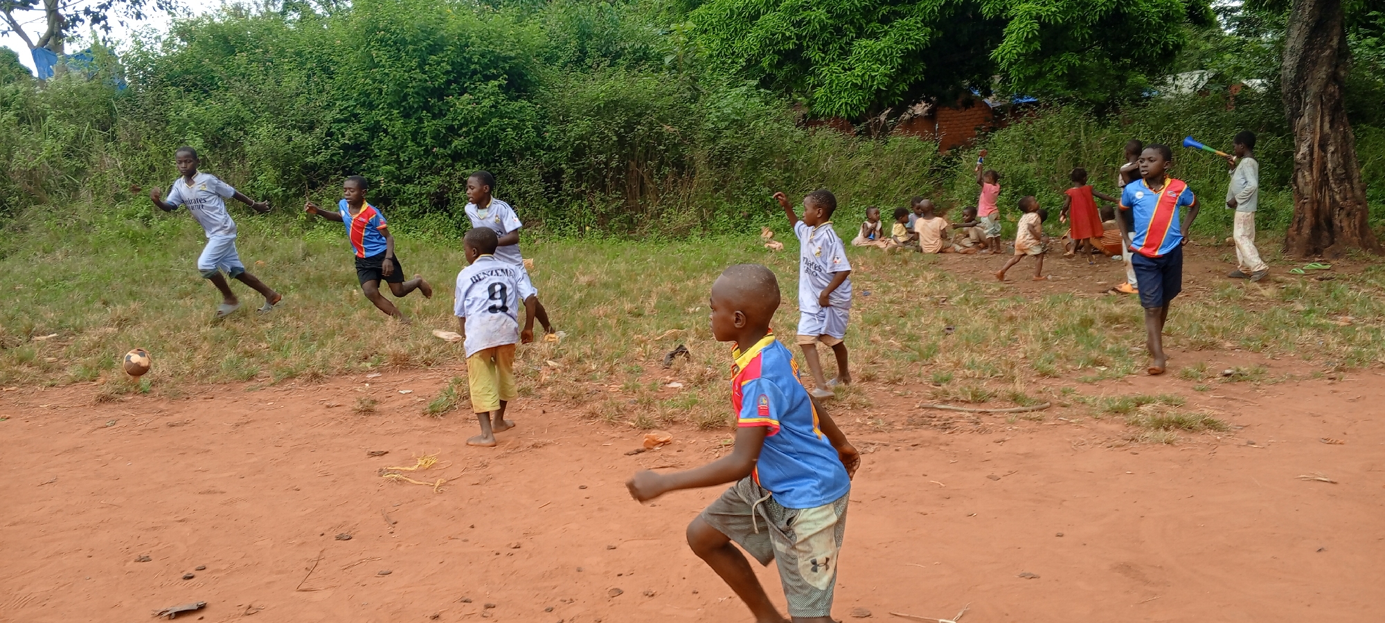 In the city of Ngalula, Democratic Republic of the Congo, a group of children are playing in an open, dirt-covered area near some trees and bushes. Most of the children are barefoot and wearing t-shirts and shorts. Some are actively playing what appears to be a game of football, with one child chasing after a soccer ball. Others are gathered in the background, sitting or standing. The setting looks like a simple and possibly impoverished neighborhood.