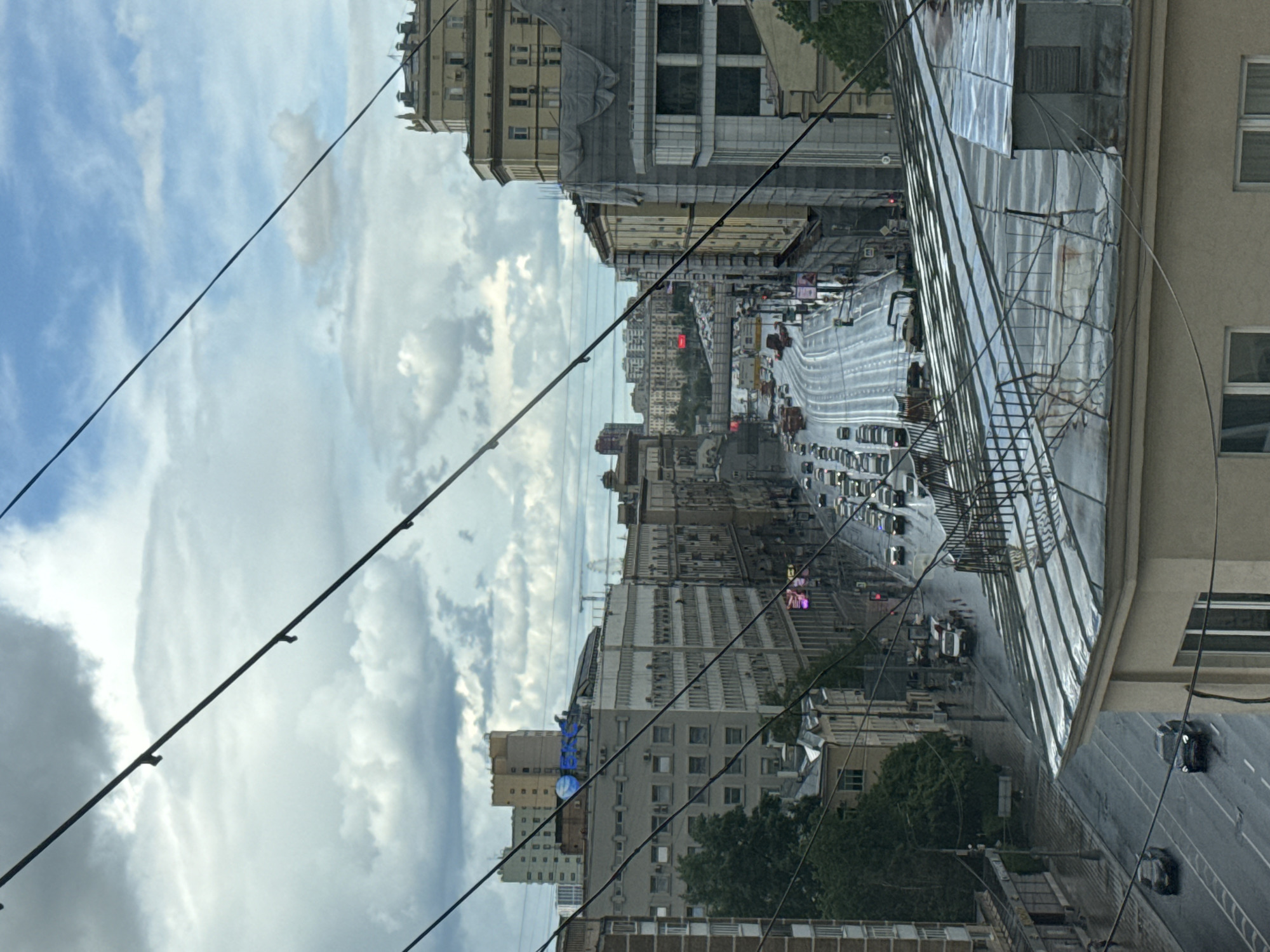 View from above looking down on a street in Moscow, Russia. The street is filled with traffic, including cars, buses, and trucks. The buildings on either side of the street are tall and imposing. The sky is cloudy. Power lines stretch across the foreground. A glass structure and buildings are visible on the right.