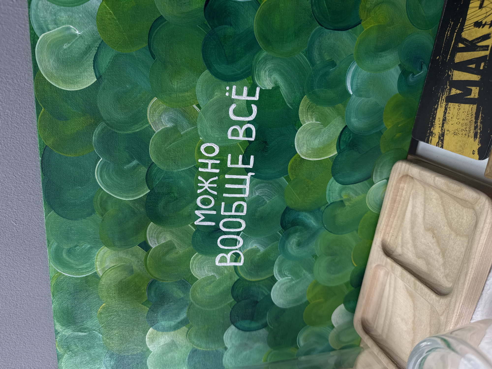 The image shows a section of a wall decorated with a painting. The painting consists of overlapping green brushstrokes resembling leaves or scales. The text "МОЖНО ВООБЩЕ ВСЁ" (meaning "Anything is possible" in Russian) is written in white on the green background. To the right of the painting are a wooden tray and the corner of a black and gold object with the letters "MAK" visible. The wall is painted gray. The location is Izhevsk, Russia, as it is suggested in the text "МОЖНО ВООБЩЕ ВСЁ", which is in Russian.