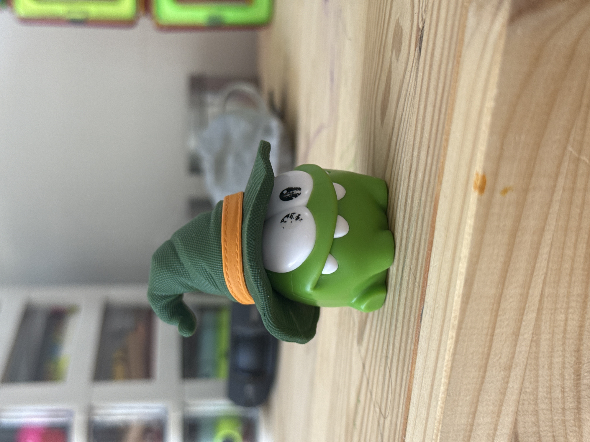 A green, frog-like toy character wearing a green witch's hat with an orange band sits on a light-colored wooden surface. The toy has large white eyes with black pupils and two small white teeth. The background is out of focus and shows shelves with various objects, including what appears to be a black electronic device and some blurred containers. A stack of bright green and red containers is visible on the far right. The image is a close-up shot of the toy, with the wooden surface dominating the foreground. The scene is indoors, likely on a table or shelf. The location is not identifiable as Lyubertsy, Russia.