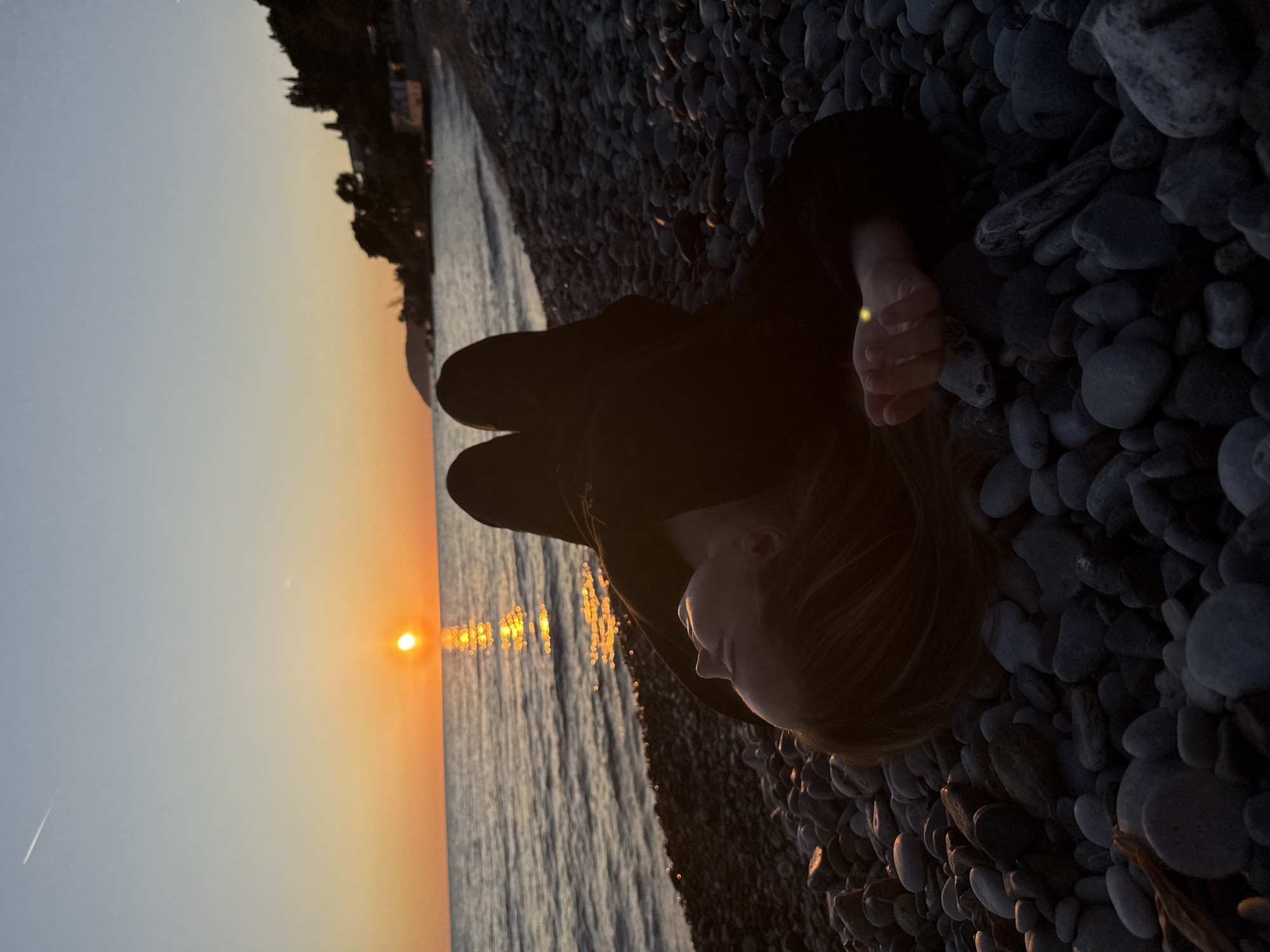 The image shows a person lying on a pebble beach at sunset. The sun is setting over the water in the background, casting a golden glow on the scene. The person is wearing dark clothing and their face is turned towards the sun. The beach is covered in small, dark pebbles. The location is likely a beach near the city of Bar, Montenegro, as the coastline is characterized by pebble beaches and sunsets over the Adriatic Sea.
