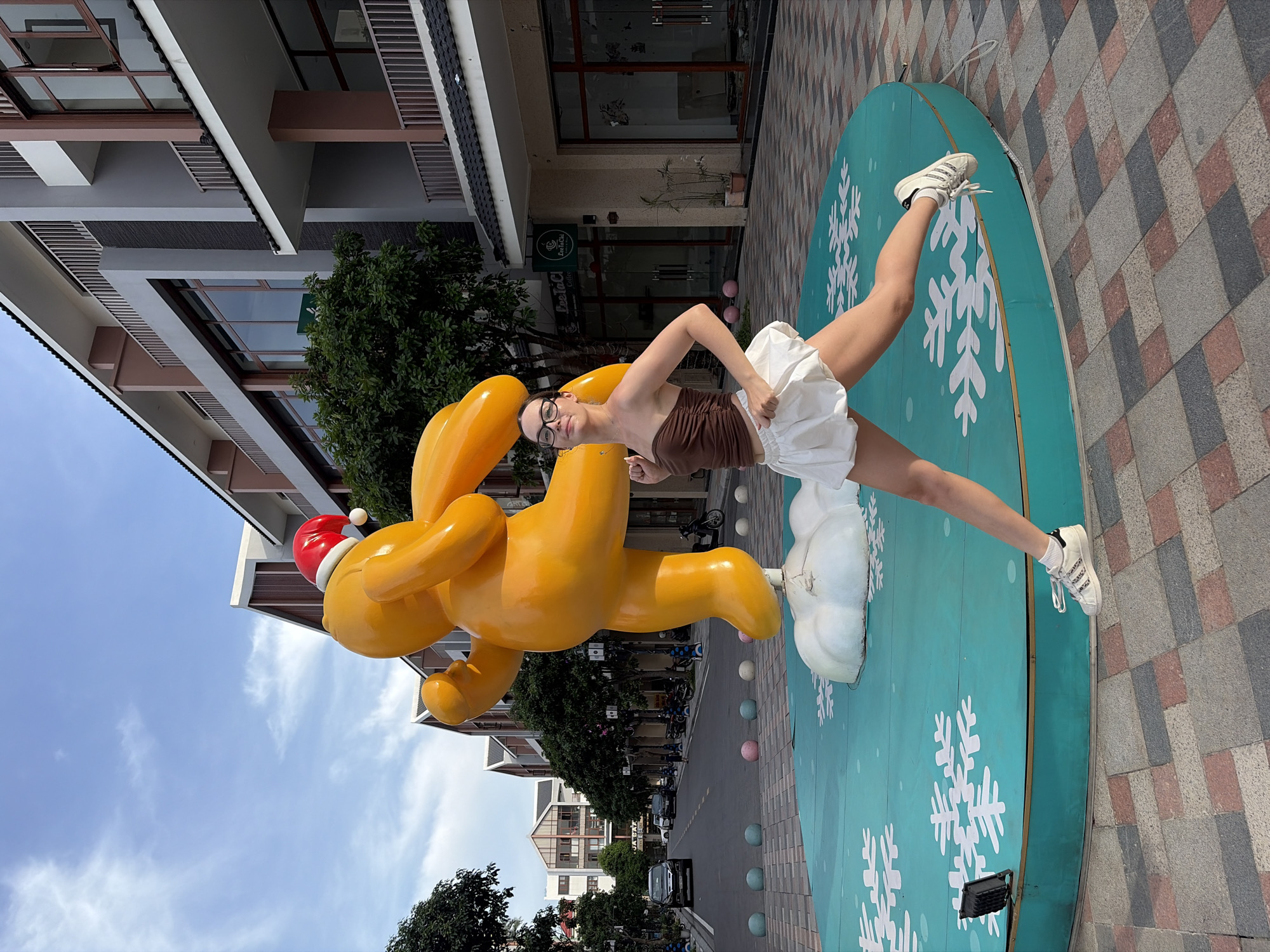 An eye-level medium shot captures a young woman with glasses mid-jump in front of a large, bright orange, inflatable sculpture of a cartoon character wearing a Santa hat. The sculpture is positioned on a teal-colored circular platform decorated with white snowflakes. The woman is wearing a brown strapless top and a white ruffled skirt, with white sneakers. Her body is angled towards the right, with her left leg extended forward and her right leg bent. The background reveals a street scene in Nghĩa Trụ Commune, Vietnam, with modern buildings lining the road. The buildings are multi-story with a mix of beige, grey, and brown facades, featuring balconies and large windows. Trees are planted along the sidewalk. Cars, motorcycles, and decorative spherical bollards are visible on the street. The sky is partly cloudy with a bright blue hue. The ground is paved with a checkered pattern of grey, pink, and beige tiles. The overall scene appears to be an outdoor public space, possibly a plaza or a pedestrian area.