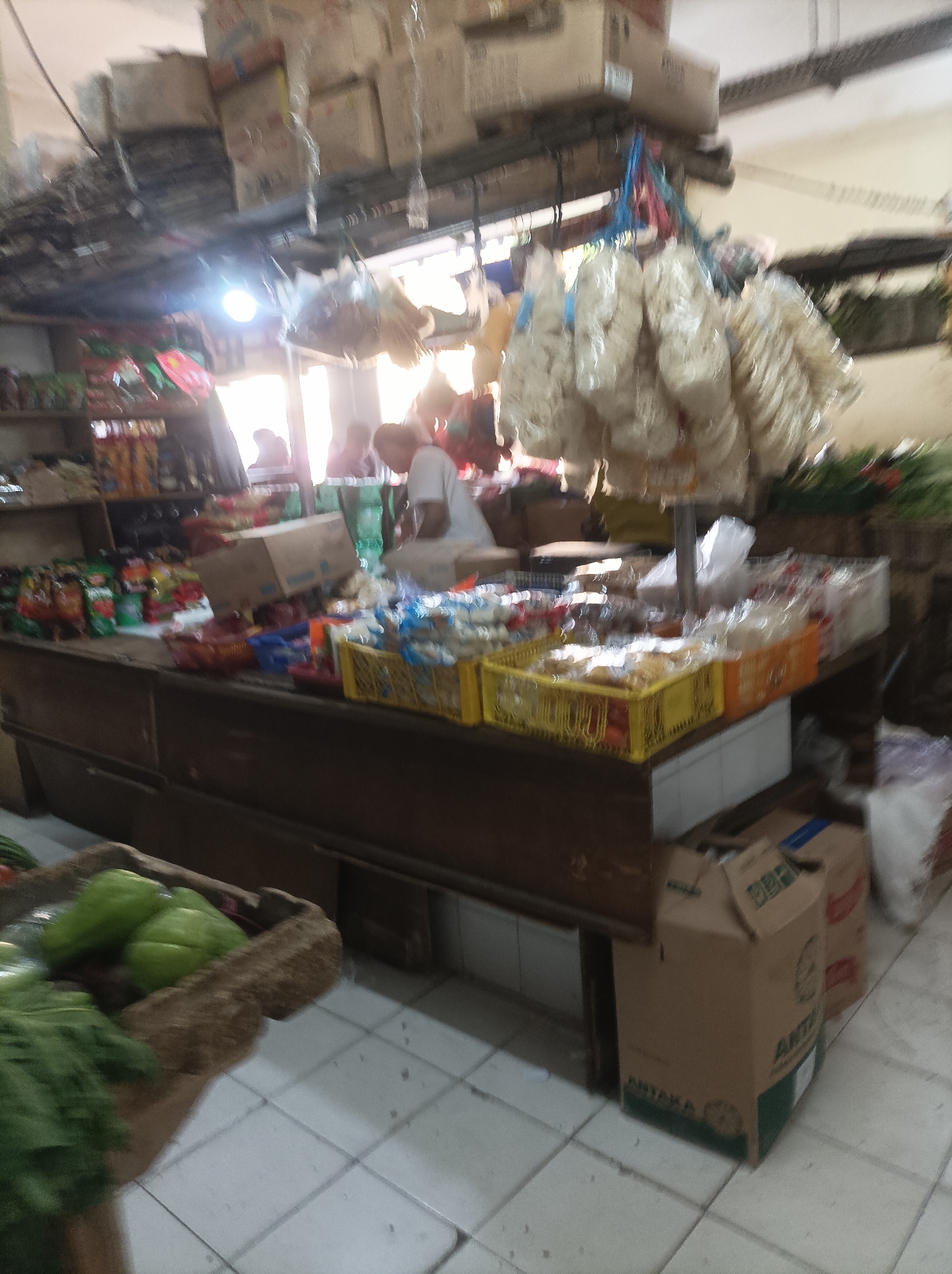 This is a blurry, eye-level, medium shot of an indoor market stall in West Jakarta, Indonesia. The stall is filled with various goods, including packaged food items, dried snacks hanging from the ceiling, and fresh produce in a woven basket in the foreground. The lighting is dim, with a bright light source visible in the upper left of the frame. Several people are present in the background, appearing to be customers or vendors. The flooring is tiled. The overall impression is one of a busy, traditional market environment.