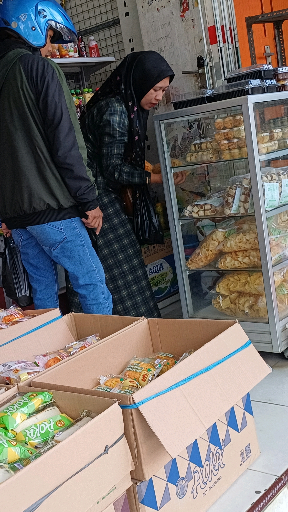 A man wearing a blue helmet and a green jacket stands to the left of a woman wearing a black hijab and a plaid dress. The woman is looking into a glass display case filled with food items. In front of the display case, several cardboard boxes are stacked. The boxes contain packaged snacks and baked goods, including bags of chips and what appear to be bread rolls. The background shows a store interior with shelves holding bottles of drinks and other merchandise. The scene is likely in a market or small shop in Pingit, Indonesia.