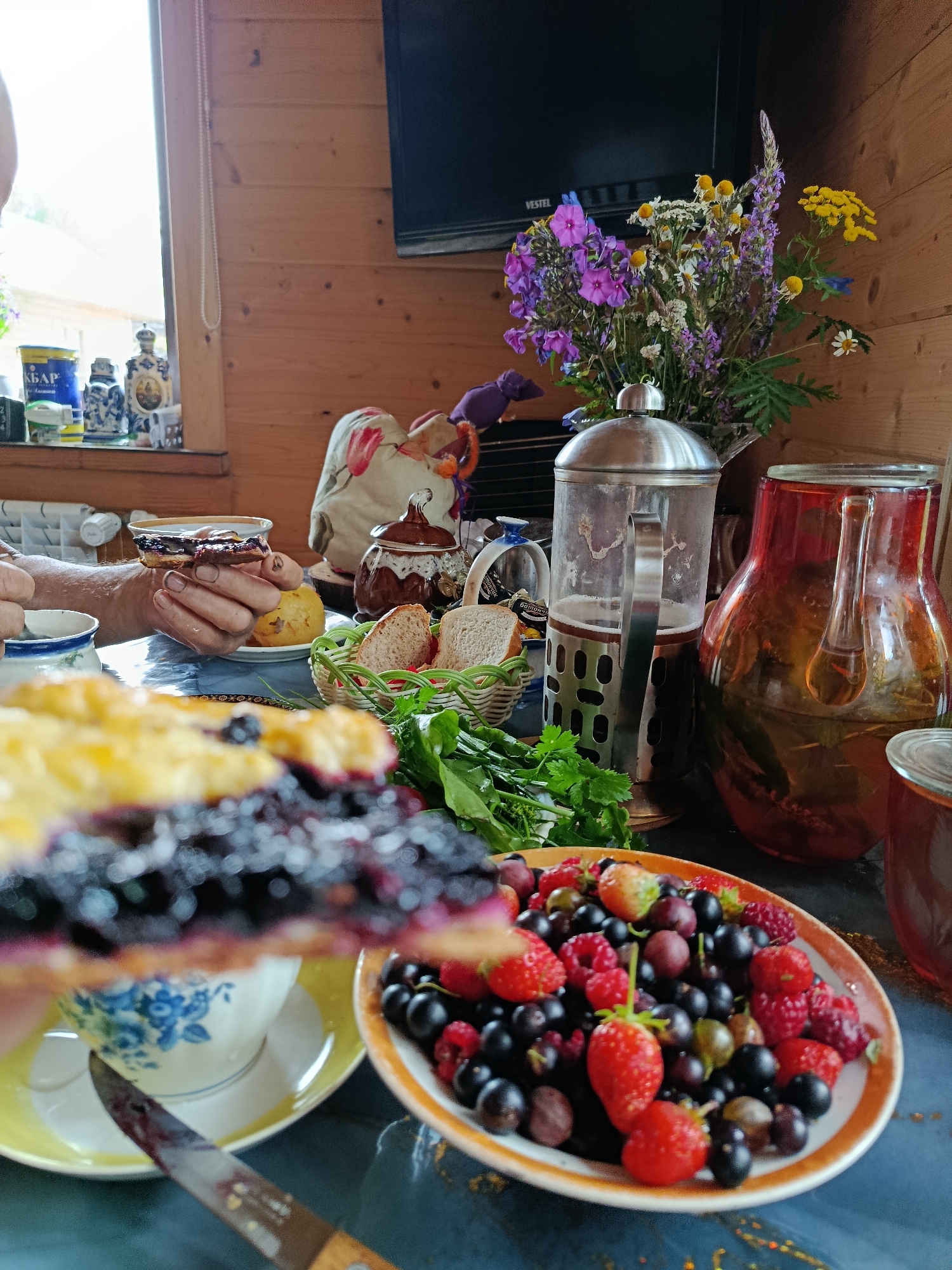 The image depicts an indoor scene of a table laden with food and drinks, likely a meal or a spread. In the foreground, a slice of pie with dark berries and a yellow topping is held by a person's hands. The pie is on a white teacup with blue floral patterns, resting on a yellow saucer. A knife with berry residue is positioned in front of the teacup. To the right of the pie, a plate is filled with an assortment of fresh berries, including strawberries, raspberries, blueberries, and gooseberries. Behind the berries, a glass French press filled with dark liquid, likely tea, is prominent. Next to it, a large, amber-colored glass pitcher contains a translucent liquid with visible herbs or leaves. In the background, a bouquet of wildflowers is arranged in a vase, and a television screen is mounted on a wooden wall. The table surface is a dark, reflective material. The environment suggests a rustic or cozy setting, possibly a kitchen or dining area with wooden paneling. The lighting is bright, coming from a window on the left side of the frame.