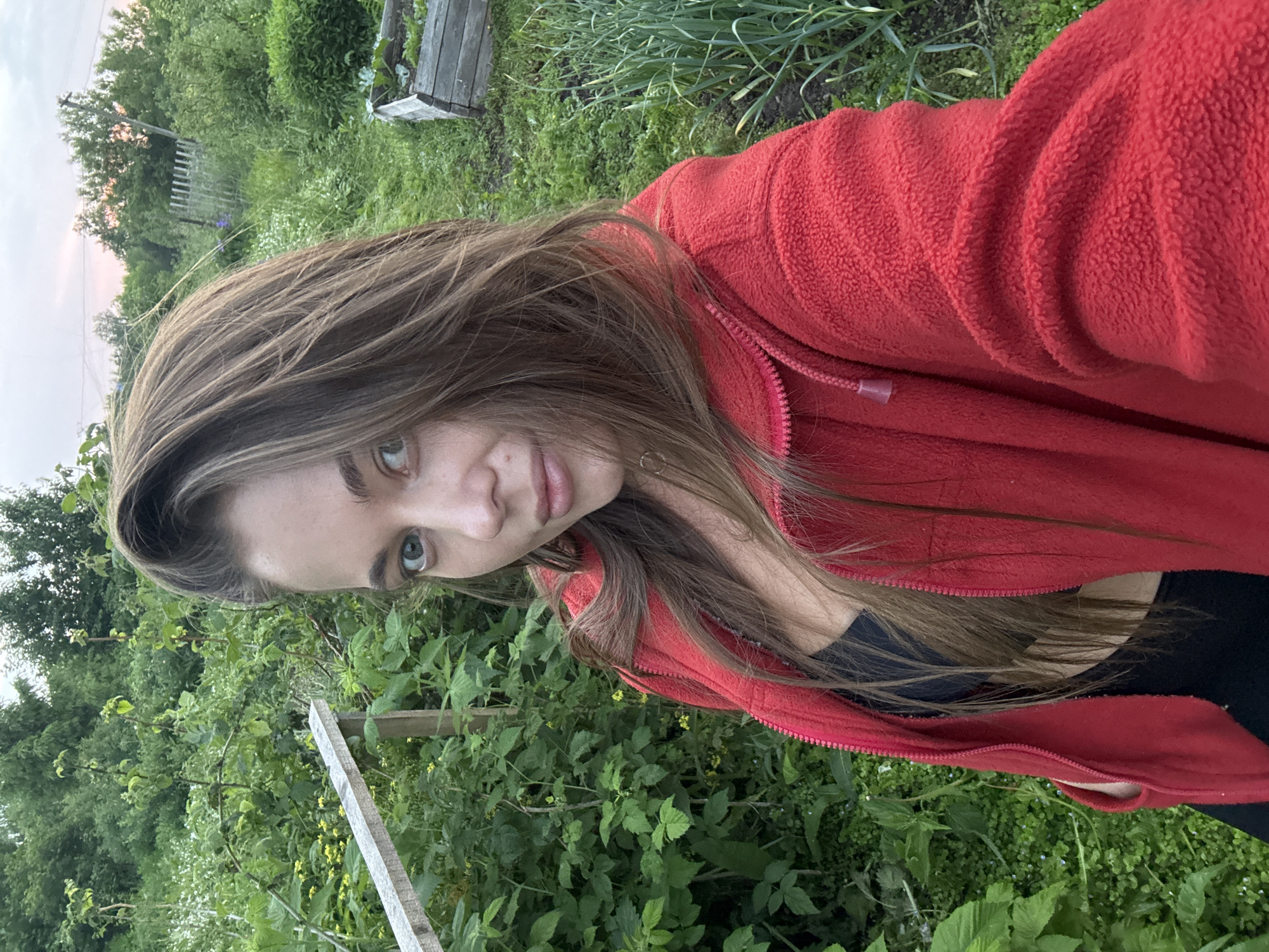 A young woman with long brown hair and blue-green eyes is pictured in a selfie taken outdoors in what appears to be a rural or garden setting. She is wearing a red fleece jacket over a black top. The background shows lush green vegetation, including plants and trees, and a wooden structure that could be a garden bed or fence. In the distance, a wooden fence and more trees are visible, with a hint of a pale pink sky suggesting dusk or dawn. The location is identified as Sulmash, Russia.