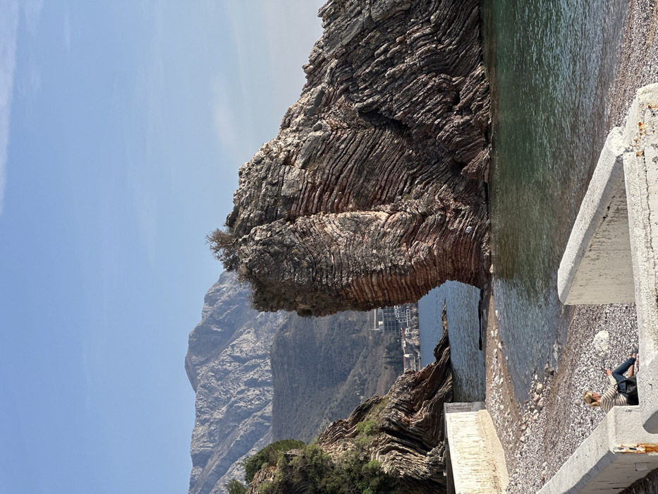 The image displays a coastal landscape in Bečići, Montenegro. The foreground features a pebble beach and sections of low, weathered concrete barriers. One adult female individual is seated on the pebbles in the lower-left quadrant, with her back to the viewer. She has blonde hair, wears a horizontally striped long-sleeved shirt and dark pants, and holds a dark handheld device. A dark shoulder bag is positioned next to her.
The mid-ground consists of calm, turquoise-green seawater extending to a prominent, layered rock formation on the right. This formation exhibits distinct reddish-brown and gray geological strata, with a small cluster of dry vegetation atop its highest point. To the left, another similarly layered rock formation, partially covered in green foliage, helps define a small cove.
In the background, several white multi-story buildings are visible at the water's edge, nestled against the foothills of large, rugged gray mountains. The sky above is clear and pale blue. The scene is illuminated by natural daylight.