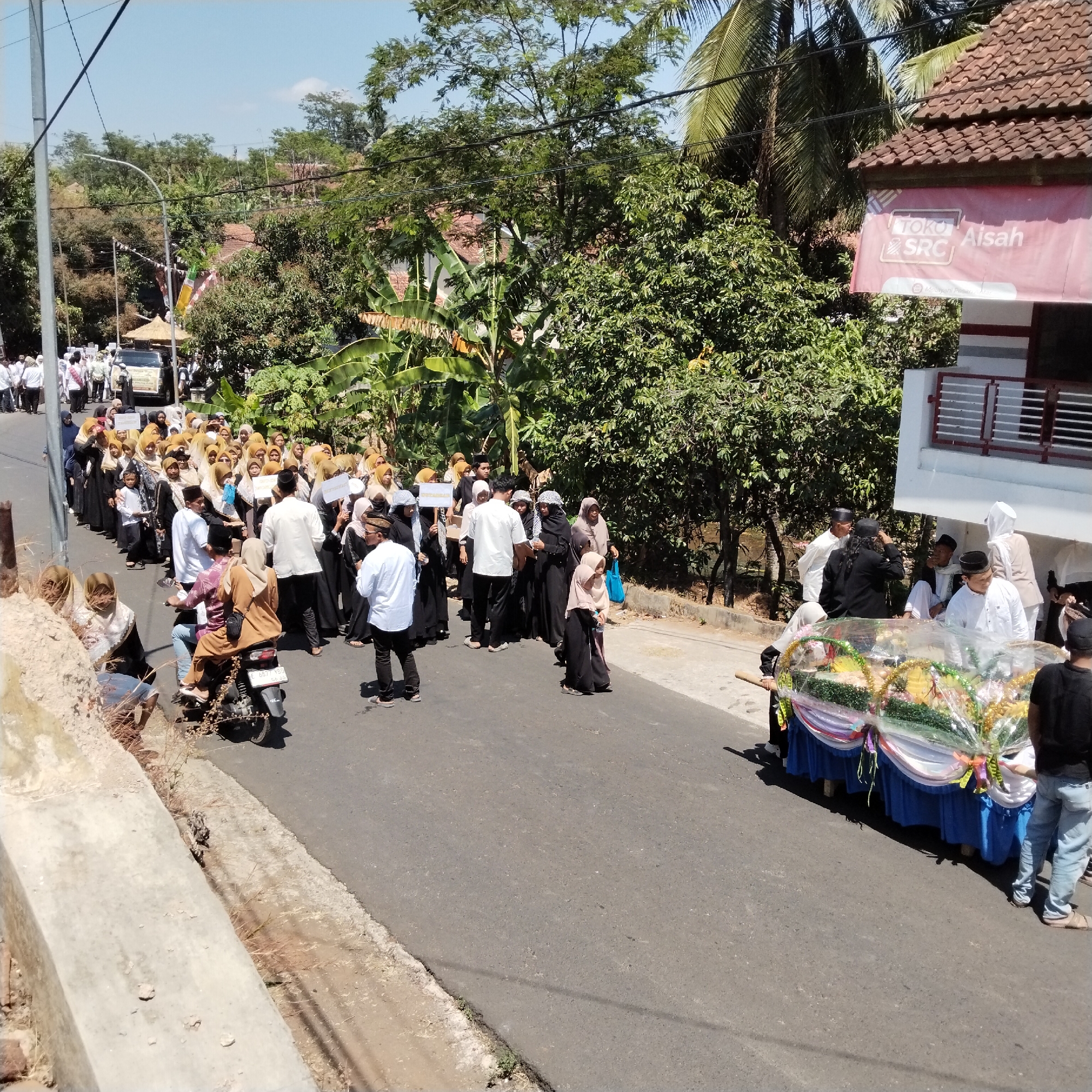 In the city of Pamulihan, Indonesia, a procession is taking place on a paved road lined with trees and houses under a clear blue sky. A group of people, many wearing white or dark religious attire and head coverings, are walking in a line. Some are holding white signs. In the foreground, a decorated float or carriage, adorned with colorful fabrics and what appears to be floral arrangements, is being pulled by several individuals. A motorcycle with two people on it is visible, and a few onlookers are standing on the side of the road. A banner with "TOKO AISAH SRC" is displayed on a building in the background.