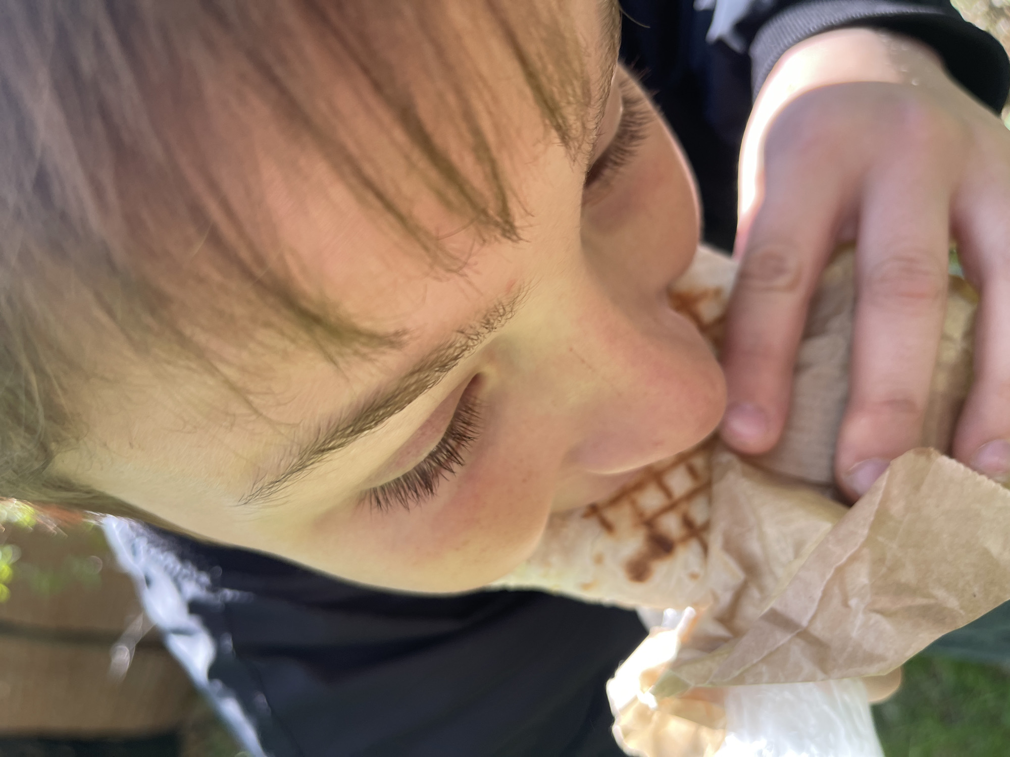 A close-up, angled shot depicts a child eating a wrap or burrito. The child has fair skin, light brown hair, and closed eyes. The wrap is held in the child's right hand and is partially encased in a brown paper wrapper. The wrap appears to contain a grilled filling. The background is out of focus, revealing hints of green foliage and a black jacket. This image likely wasn't taken in Минино, Russia, as the location cannot be determined from this image.