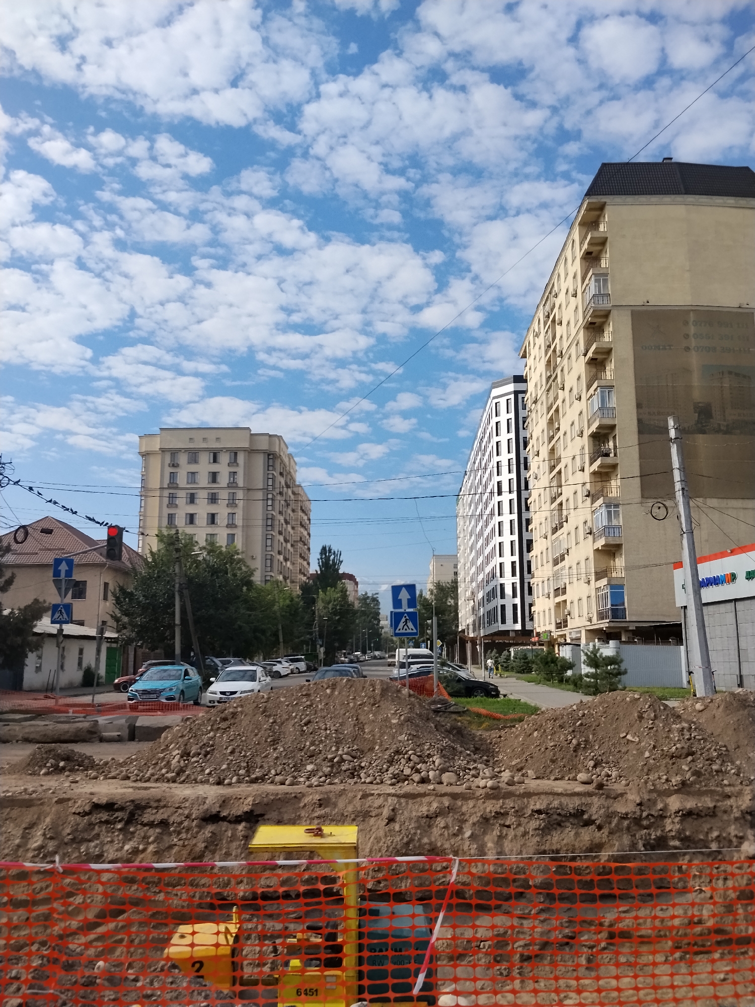 This is a photograph taken from street level, looking up towards apartment buildings and a street scene. In the foreground, construction is evident, with piles of dirt and rubble, and orange safety fencing partially obscuring some yellow construction equipment. Beyond the construction, a street is visible with several cars parked and driving. Traffic lights and pedestrian crossing signs are present. Two tall apartment buildings dominate the middle ground, one a light yellow color with balconies and the other a more modern white and black building. The sky above is a bright blue with scattered white clouds. The overall impression is an urban environment undergoing construction, with residential buildings and daily life on the street continuing. Based on the context of the image and common landmarks, this scene is likely located in Bishkek, Kyrgyzstan.