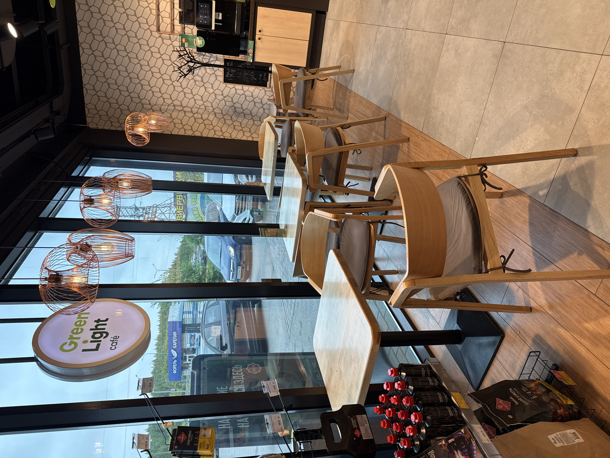 The image captures the interior of "Green Light Cafe" in Medovoye, Russia, featuring a row of tables and chairs arranged along a window. The "Green Light Cafe" sign is visible through the window. Outside the window, a street scene includes cars and a billboard advertising "Forel" (trout) from Karelia. Shelves beneath the tables display products like charcoal briquettes and bottles with red caps. The interior design includes modern lighting fixtures and patterned wallpaper. The flooring is a combination of tile and wood.