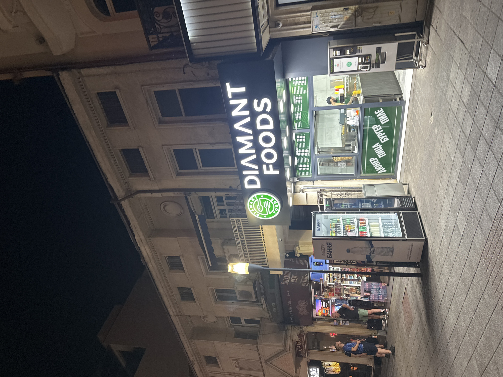 A night time exterior street view shows a food establishment called "DIAMANT FOODS". The establishment features a bright neon sign with the name in white letters against a dark background, with a green circular logo to the left. Below the sign is a glass-enclosed counter where food is being prepared, with a vertical spit of meat visible. The menu board above the counter displays green text on a black background. In front of the establishment, a tall, illuminated street lamp stands on the sidewalk. To the right of the food establishment, a white vending machine with a dark screen and buttons is present. Further to the left, other shops are visible, including one with a sign reading "CAIRO IRISH TRUE". Two men are standing on the sidewalk to the left of the scene. The ground is paved with square tiles. The overall scene depicts a commercial street at night.