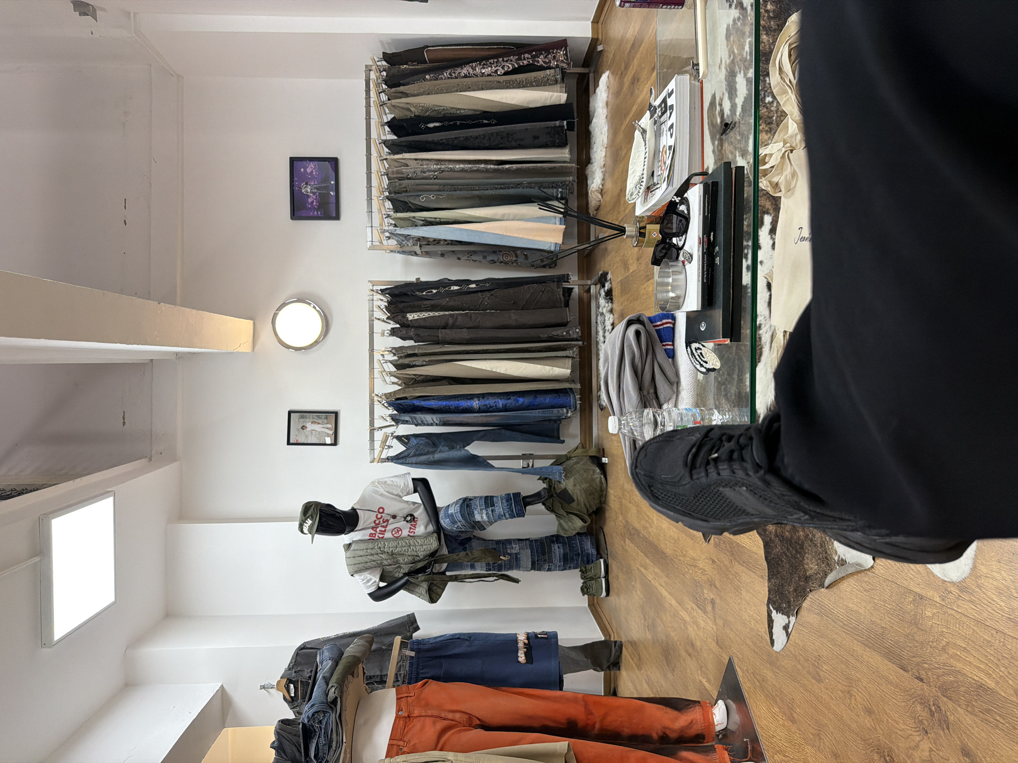The image depicts the interior of a retail clothing store, Bacchus Kills, located at 75 Berwick Street, Soho, London, United Kingdom. The scene is viewed from a low perspective, showing the extended legs and feet of an individual wearing black trousers and black sneakers.
In the immediate foreground, a clear glass coffee table is visible, holding a plastic water bottle (Volvic), a stack of folded grey knitwear, a fragrance diffuser with black reeds, a small silver bowl, black sunglasses, a silver ring, and several stacked books, one partially titled "JAPAN." Beneath the table, on the light brown wooden floor, a partial brown and white cowhide rug is laid.
The midground showcases the store's merchandise and fixtures. To the left, a partial mannequin displays orange trousers with a black dye effect and blue shorts. Center-left, a black headless mannequin is dressed in an olive green cap, a white t-shirt featuring "BACCO KILLS" and "STARS" text, and blue patchwork jeans. An olive green quilted jacket is draped over its left shoulder, and its left foot rests on a large olive green duffel bag. A pair of olive green sneakers are visible on the floor near its feet. Two freestanding metal clothing racks are positioned against the white back wall, laden with numerous pairs of trousers and other garments in varying dark and muted colors. Small white faux fur rugs are placed beneath each clothing rack.
The background consists of plain white walls. On the left wall, a small framed picture of a person in white clothing is mounted. On the back wall, a circular light fixture is present to the left, and a framed monitor or picture displaying a person on a purple-lit stage is to the right. The white ceiling features a large rectangular LED light panel and visible structural beams.