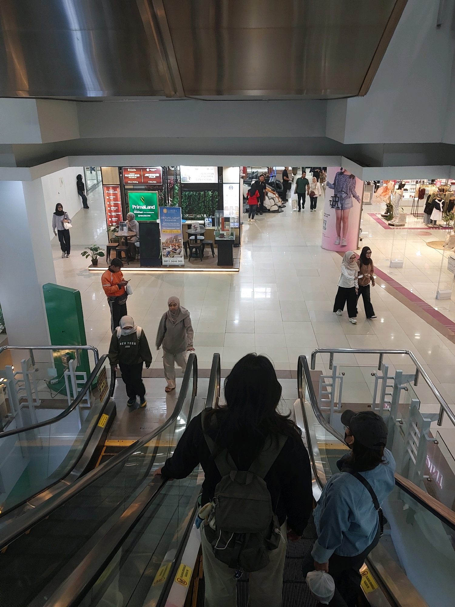 The image captures an indoor scene, likely a shopping mall, with people navigating an escalator. In the foreground, two individuals are descending on the escalator, their backs to the camera. One is wearing a black hoodie and a backpack, while the other sports a blue jacket and a baseball cap. In the mid-ground, several people are visible, some walking and others seated at tables in what appears to be a cafe or food court area. Signage for businesses such as "PRIMALAND" is present. The environment is brightly lit with a tiled floor and modern architecture, suggesting a contemporary commercial space. The context implies a public area, possibly in the city of Malang, Indonesia, given the presence of Indonesian signage and common mall features.