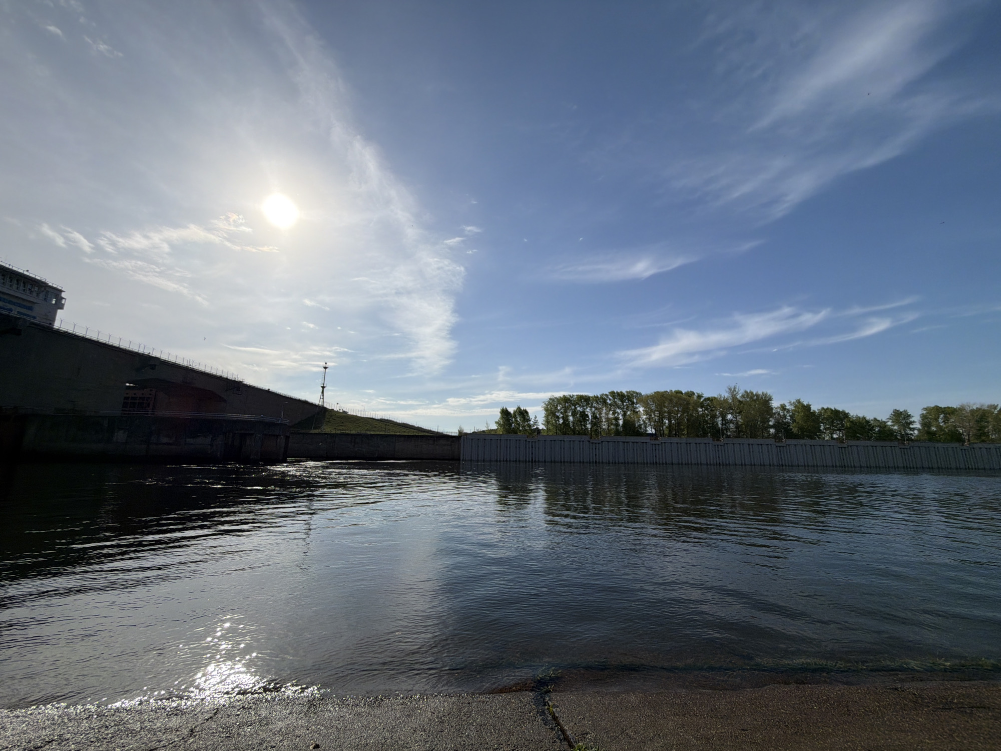 The image shows a view of the Kama River in the city of Chaikovsky, Perm Krai, Russia. The river is in the foreground with reflective water. A large concrete structure, possibly a dam or part of a power plant, is on the left side of the image. On the opposite side of the river there is a long, man-made structure and trees in the background. The sky is blue with scattered clouds, and the sun is visible in the upper left corner of the image. The perspective is from a low angle, likely near the riverbank.