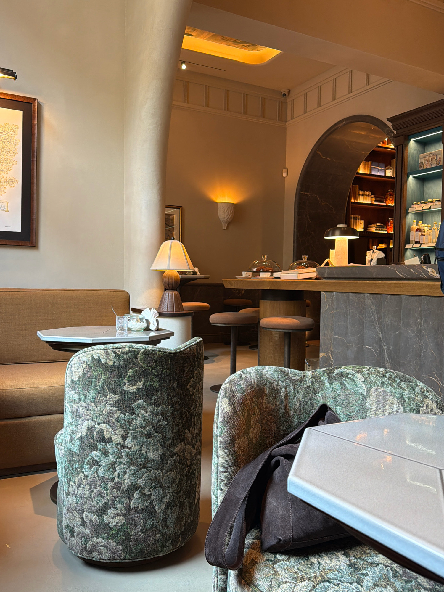 The image depicts the interior of a cafe, likely in Milan, Italy, characterized by its sophisticated and modern decor. In the foreground, a plush armchair upholstered in a vintage floral fabric is partially visible, with a dark brown suede bag resting on it and a sleek, light-colored table in front. To the left, a sofa with a similar neutral upholstery anchors the seating area, accompanied by a hexagonal table featuring a small vase with a white flower and a glass.
The midground reveals a bar area, demarcated by an arched opening with dark, textured walls. Behind the counter, which has a wooden top and a marble-patterned base, a display cabinet filled with bottles and books is illuminated by blue LED lights. Two modern desk lamps and a glass cloche covering food items are visible on the counter. Several bar stools with brown seats and dark legs are placed around the counter.
The background shows a wall with a textured finish and a modern sconce emitting a warm light. Further back, recessed lighting illuminates the ceiling, and a security camera is subtly placed on the wall. The overall ambiance is warm and inviting, suggesting a chic establishment with attention to detail in its design and furnishings. The presence of a framed artwork on the left wall adds a touch of elegance to the space.