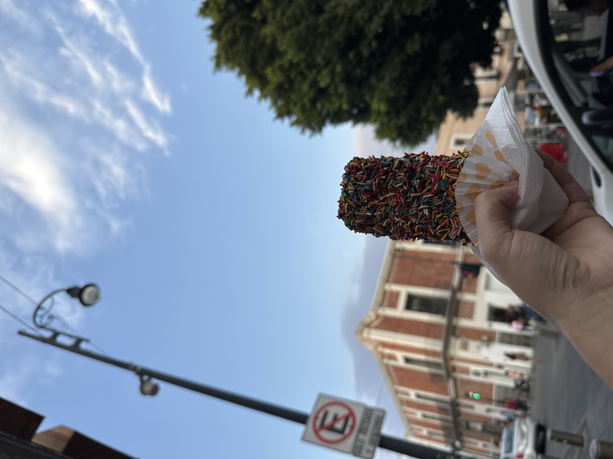 A person is holding a pastry covered in colorful sprinkles in front of a street scene. The pastry is held in a white and light brown paper wrapping. Behind the pastry, there is a building with a brick facade and white trim, several windows, and visible street activity, including cars and pedestrians. The sky is blue with scattered clouds and a tree with dense green foliage is visible in the upper part of the frame. A street lamp and a traffic sign are in the foreground. The location is city of Unknown, Unknown.