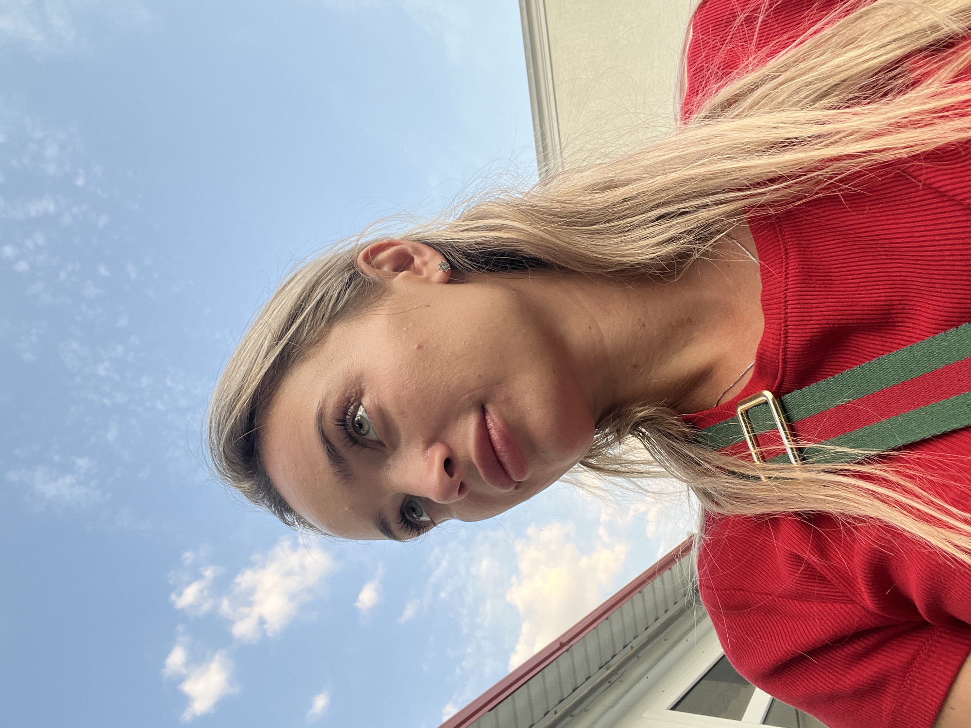 The image shows a close-up of a young woman's face, looking upwards towards a blue sky with scattered clouds. She has blonde hair, green eyes, and fair skin. She is wearing a red shirt with a green and red striped strap across her shoulder. In the background, there is a hint of a building with a red roof on the left and a light-colored wall on the right. The lighting suggests it is daytime. The photo appears to be taken outdoors in a setting that could be a residential or commercial area.