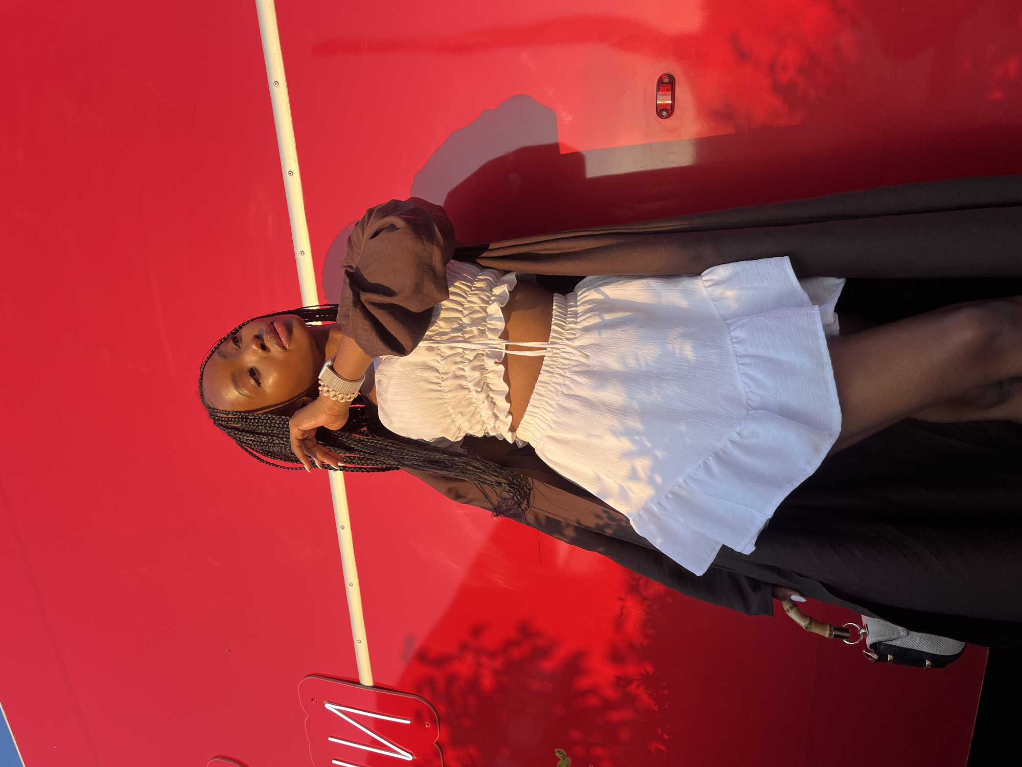 A young Black woman is posing against a bright red, wall-like structure, possibly a vehicle or building exterior. She is wearing a white ruffled crop top and a white ruffled mini-skirt, with a dark brown duster coat draped over her shoulders. Her hair is styled in long braids. She is accessorized with a bracelet on her left wrist and is holding a small purse with a bamboo handle. The background features a horizontal white stripe and a shadow of what appears to be foliage. The lighting suggests it is either sunrise or sunset, casting warm light and distinct shadows on the red surface. The image is shot in a portrait orientation. There is no discernible information in the image that can identify its location within the city of Kazan, Russia.