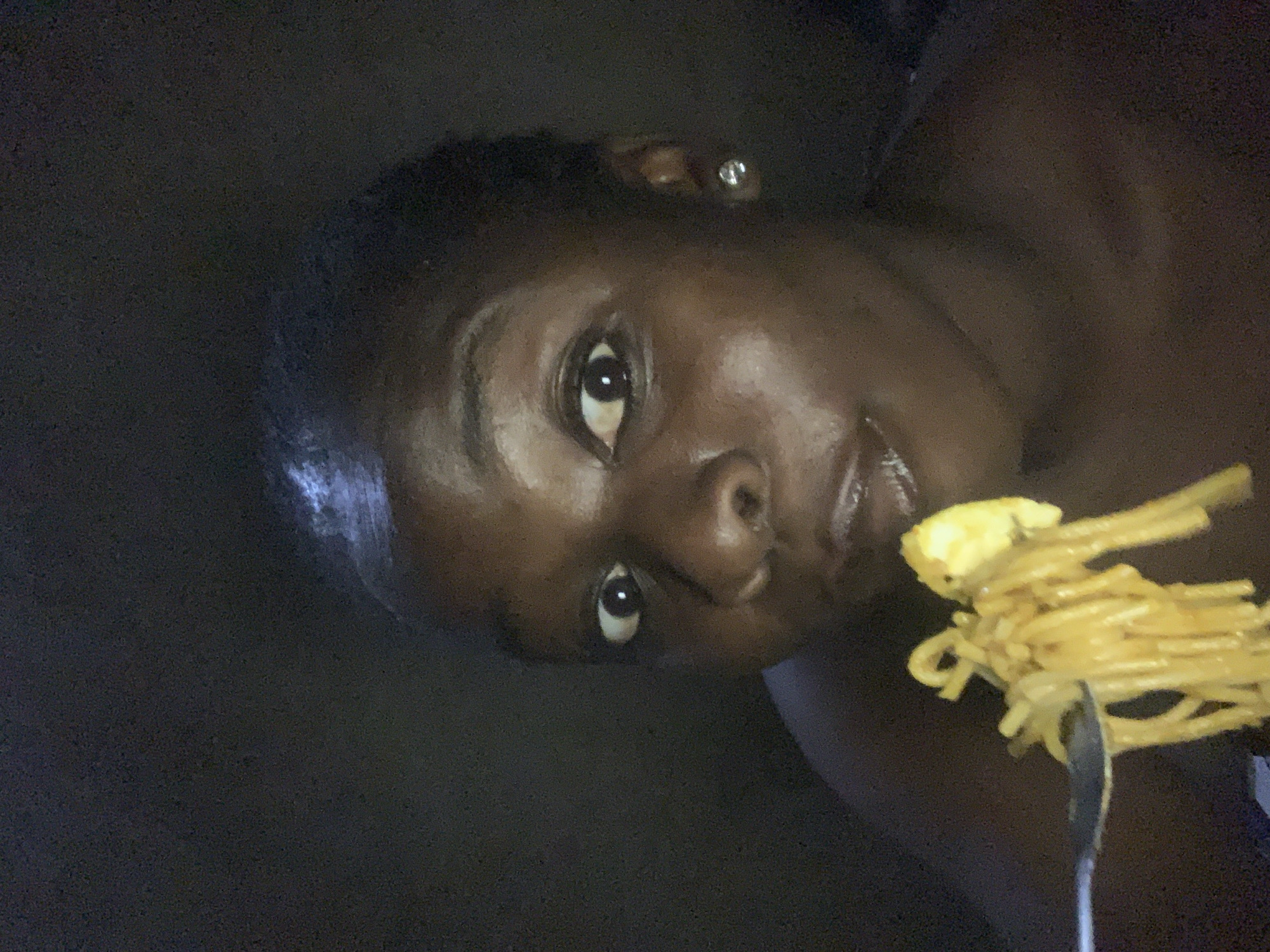 In the city of Agban, Nigeria, a young Black woman with dark skin and short hair is captured in a dimly lit, close-up selfie. She is looking directly at the camera with wide, curious eyes, her mouth slightly open as she brings a forkful of spaghetti to her lips. The spaghetti appears to be cooked and coated in a sauce, with a piece of what looks like scrambled egg on the fork as well. She is wearing a small, sparkling stud earring in her visible ear. The background is dark and indistinct, suggesting an indoor setting at night. The lighting is minimal, highlighting her face and the food.