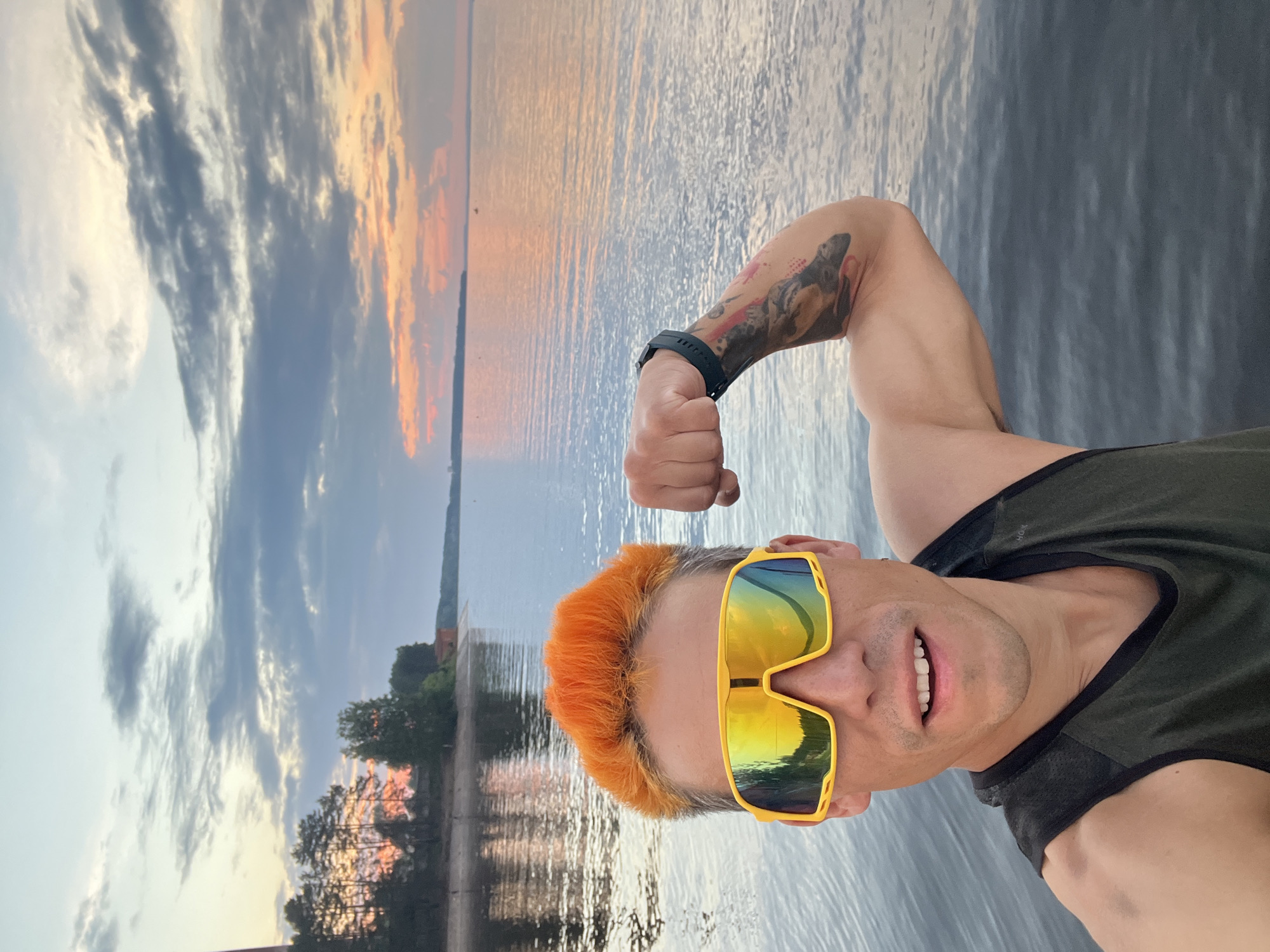 A man with bright orange hair and yellow sunglasses is posing for a selfie by the water at sunset. He is flexing his bicep, revealing tattoos on his arm. The background shows a body of water, a shoreline with trees and a building, and a colorful sky with clouds. The location is the Kama River in Chaikovsky, Russia.