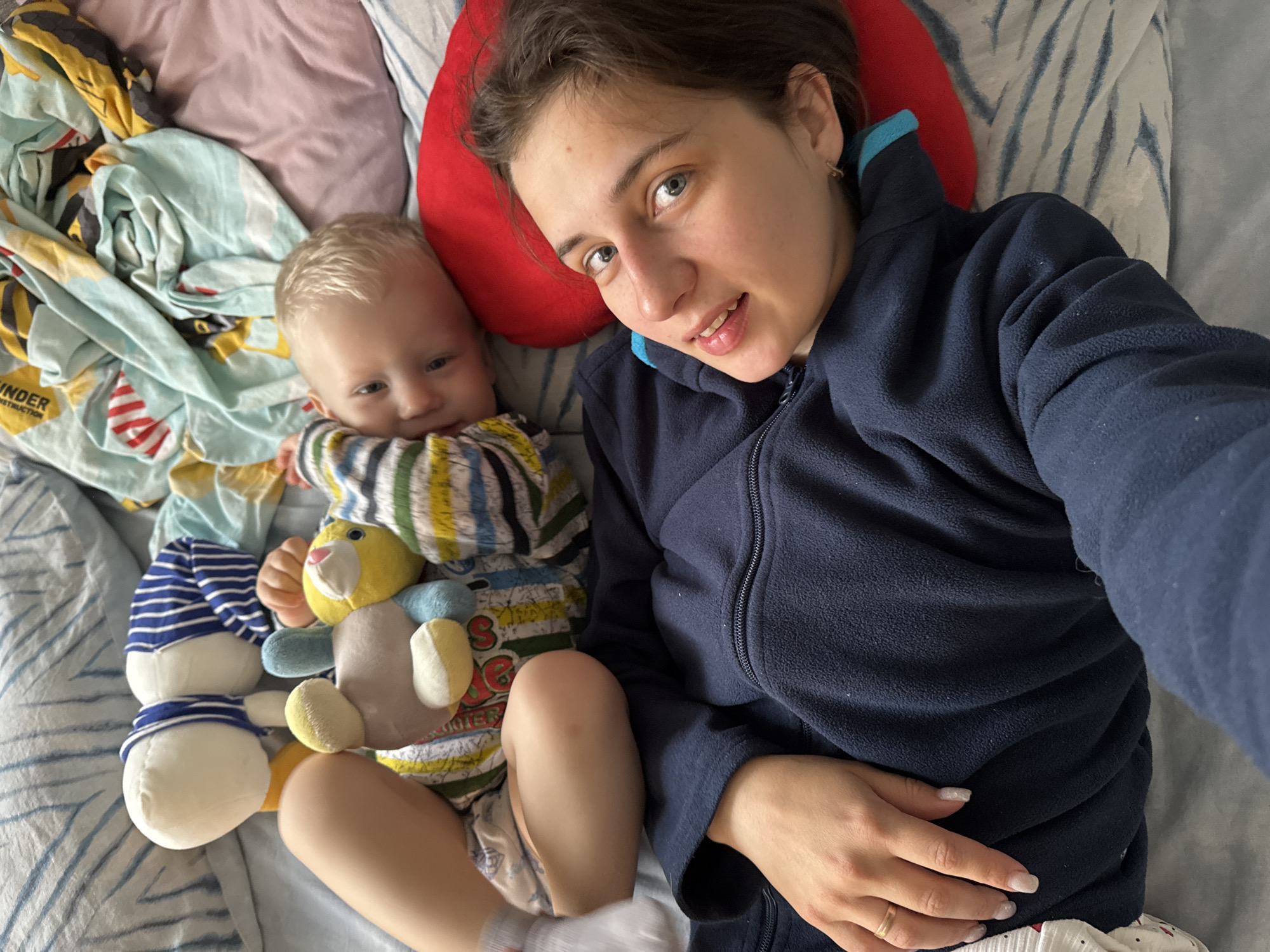 The photograph shows a woman and a baby lying on a bed. The woman, positioned on a red pillow, is wearing a navy blue zippered fleece jacket. Her hand rests on her stomach. Beside her, the baby, clad in a striped top and diaper, lies clutching a plush toy. The bed is covered in a patterned blue and white sheet, with additional patterned fabrics near the baby. The environment appears to be indoors. There is no information present that confirms that the location is Chaikovsky, Russia.