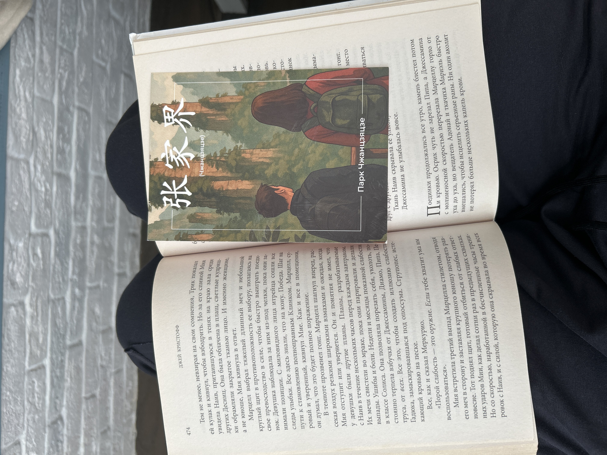This media file shows an open book with a bookmark placed on one of its pages. The bookmark features an illustration of two people, a woman and a man, with backpacks, standing in a natural landscape with trees and mountains. The book itself displays text in Russian, with Chinese characters also visible. The title "张家界" (Zhangjiajie) and "Парк Чжанцзяцзе" (Zhangjiajie Park) are prominent on the bookmark, suggesting the illustration is related to this location.
The text on the open page appears to be a narrative, possibly a story or novel, with character names like "Джей Кристофф," "Мия," "Марцелл," "Наив," "Джессамина," and "Меркурио" mentioned. The passage describes a scene of combat or a duel, with mentions of swords, shields, and injuries. There are also references to training, strategy, and deception, suggesting a plot involving conflict and hidden motives. The page number "474" is visible at the bottom left.
The overall scene is framed by a person's dark clothing, suggesting the book and bookmark are being held or displayed against this background. The lighting appears to be from an overhead source, casting some shadows. The image captures a moment of reading or presentation of literary material, with a focus on the visual elements of the bookmark and the textual content of the book.