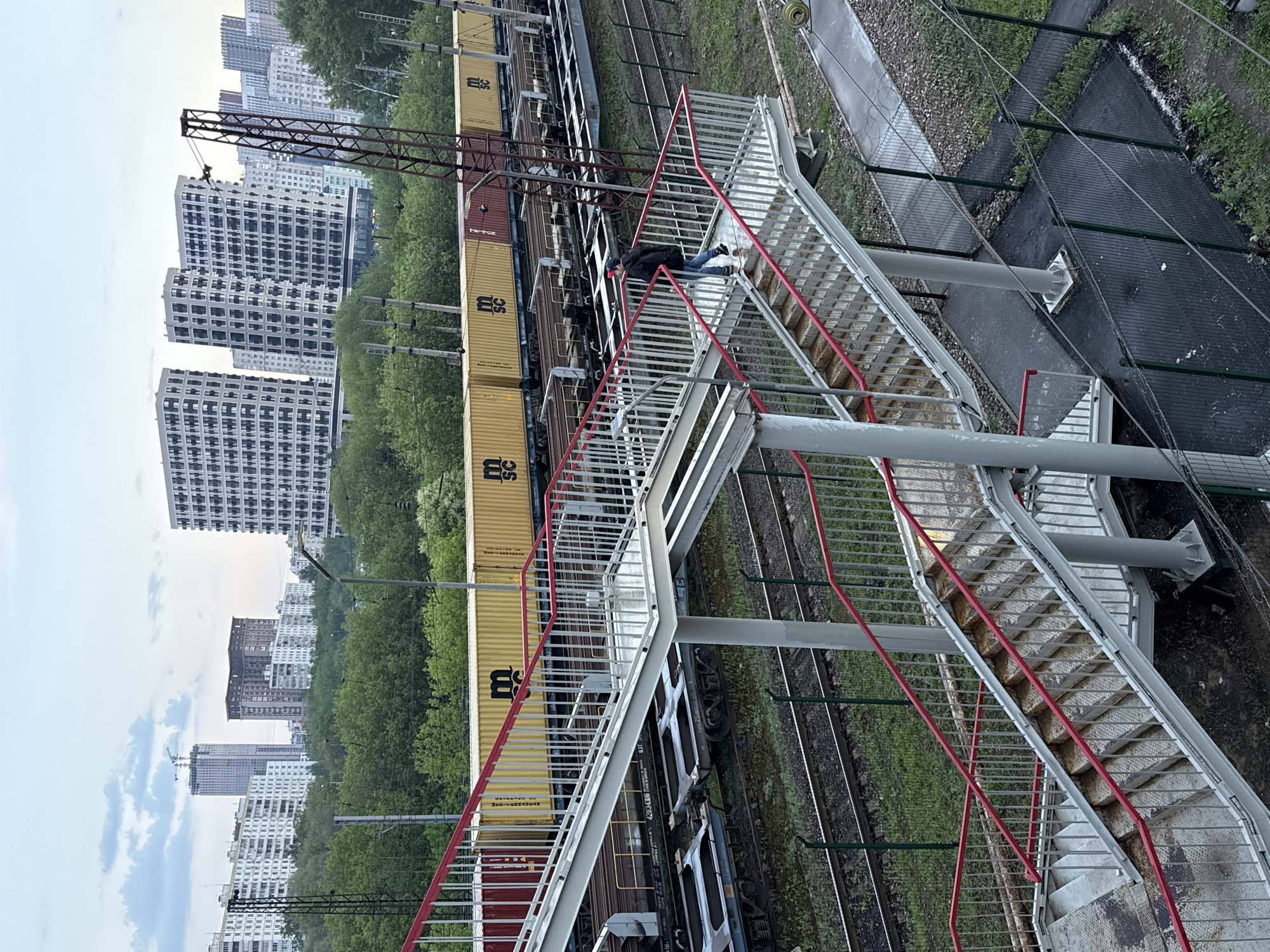 The image shows a train crossing under a pedestrian overpass, with several high-rise apartment buildings in the background, likely located in Moscow, Russia. A person wearing a dark jacket, blue jeans, and a red baseball cap is lying on the overpass between sets of stairs. The train consists of several cars and a crane is visible overhead. The overpass is constructed of metal stairs with red railings. The landscape includes train tracks and grass.