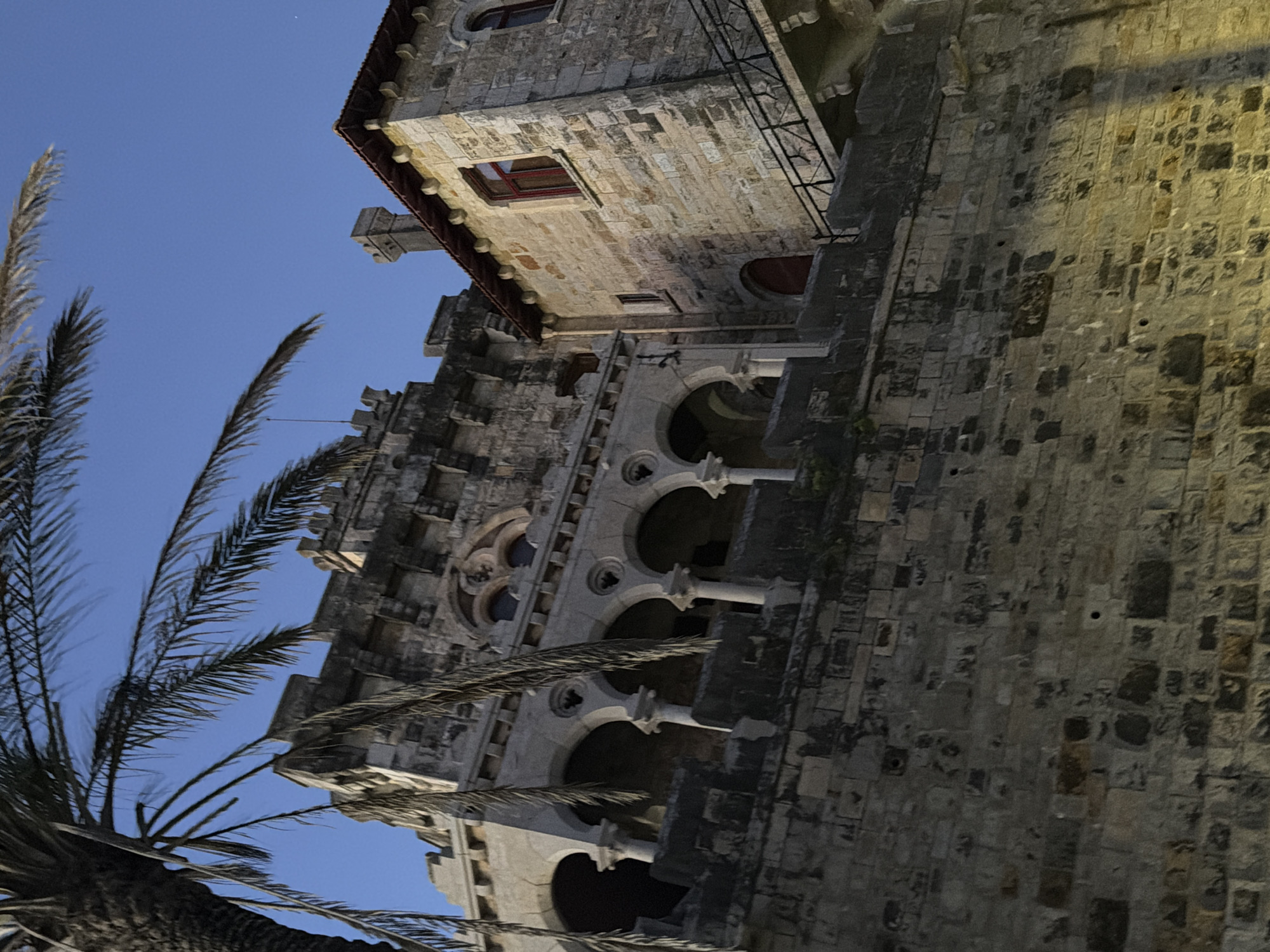 The image depicts a view of the Citadel of Cascais, Portugal, at dusk. A palm tree frond extends into the upper left corner of the frame, partially obscuring a section of the stone building. The Citadel itself is constructed of weathered stone blocks. The main building in the foreground features a series of arched openings supported by columns, above a rougher stone wall. To the right, a smaller, more rectangular stone structure with red-framed windows and a tiled roof is visible, connected by a walkway or bridge with a black metal railing. The sky above is a deep blue, indicating the late hour. The lighting suggests artificial illumination, casting shadows on the stonework. There are no people or animals visible in the scene.